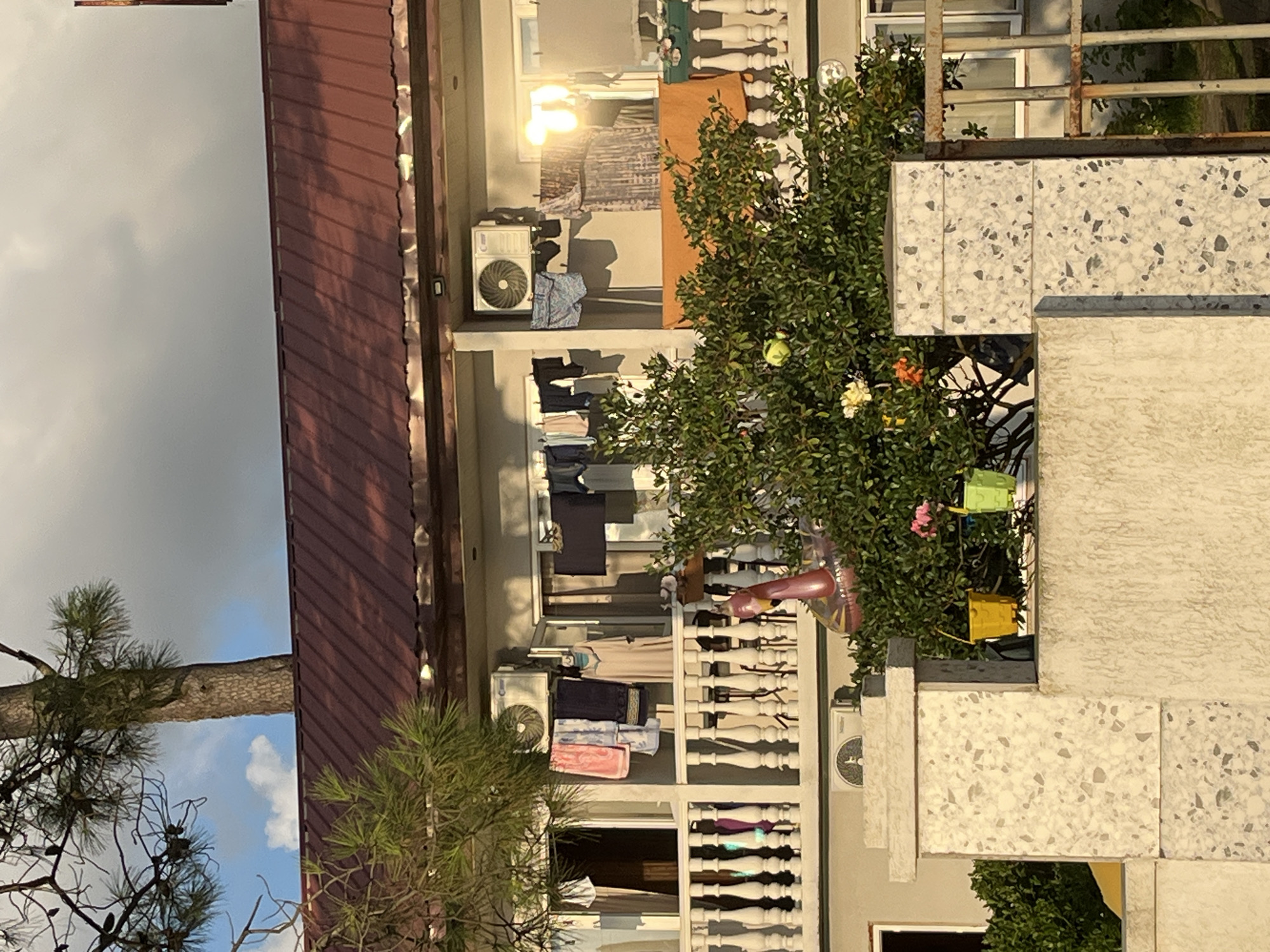 In Ureki, Georgia, a multi-story building features a red metal roof and balconies adorned with white balustrades. Laundry is hung out to dry on clotheslines on the balconies, including towels, shirts, and other garments. Air conditioning units are mounted on the exterior walls. A lush green tree with visible fruit, possibly apples, grows in front of the building. There is also an inflatable pink flamingo float visible amongst the foliage. In the foreground, a stone pillar with a speckled pattern stands near a textured wall. The sky above is a mix of blue and grey clouds, with sunlight casting warm tones on the scene.