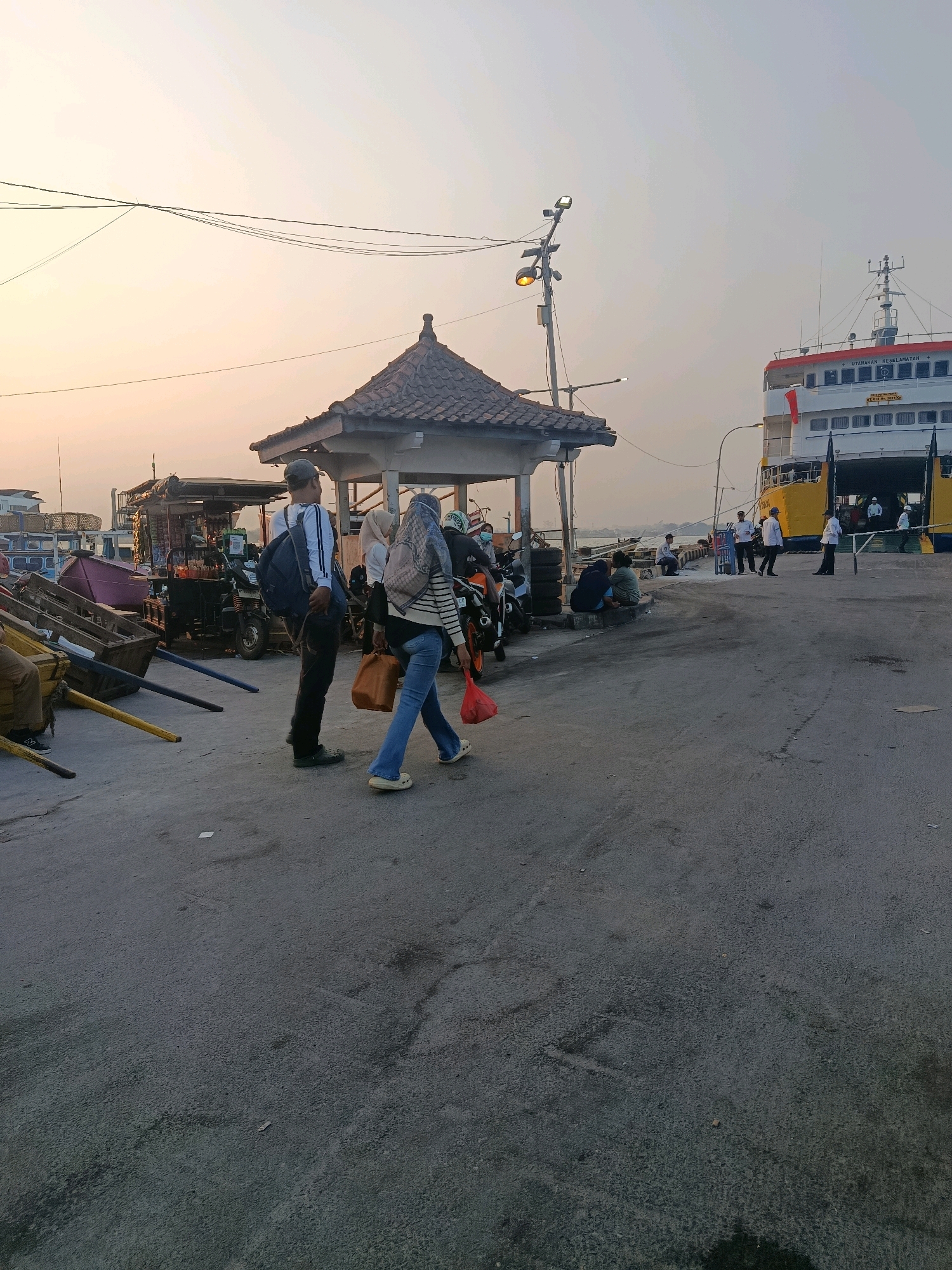 The image captures a scene at the Jepara port in Indonesia during what appears to be late afternoon or early evening. A large ferry, painted yellow and white, is docked on the right side of the frame, its ramp lowered. Several people, some in white uniforms, are visible on the ferry and near its ramp.
In the foreground and mid-ground, a paved area, likely the port's ground, is visible. A covered pavilion with a tiled roof stands to the left of center. Several people are walking or standing around this area. Two individuals are walking towards the right, one carrying a red bag and another holding a brown bag. To their left, a man with a backpack is standing, facing away from the camera. Various objects, including what look like carts and possibly market stalls, are present in the background to the left. Motorbikes are also visible. The sky is clear with a pale orange hue, indicating either sunrise or sunset. The overall atmosphere suggests a busy port environment with ongoing passenger and vehicle activity.