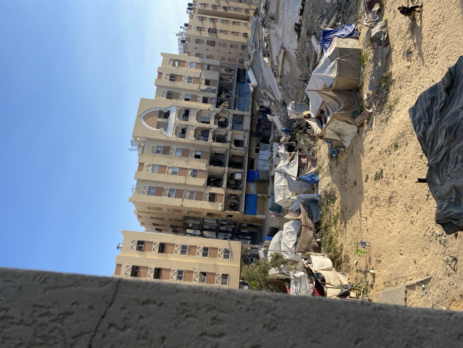 The image captures a scene of urban destruction and makeshift living under a clear, bright blue sky. The central focus is a large, multi-story apartment building, light-colored (likely beige or yellow), exhibiting extensive damage. Many windows are either completely missing, broken, or boarded up, and the facade is heavily pockmarked with numerous small holes, suggesting bullet or shrapnel damage. Several sections of the building appear partially gutted or collapsed, exposing the interiors of multiple floors. Large water tanks are visible on the building's roof. A distinctive arched architectural detail is present on one of its higher facades.
To the left and slightly in front of this severely damaged building, a second, shorter building is visible, also showing signs of damage, primarily broken windows.
The ground area in front of and surrounding these buildings is a landscape of rubble, dirt, and debris, including fragments of concrete and twisted metal. Amidst this devastation, numerous makeshift shelters, or tents, are clustered together. These shelters are constructed from various materials such as tarpaulins, sheets of fabric, and possibly some corrugated metal, suggesting a temporary encampment for displaced individuals.
A dark-colored dog is seen walking on the sandy ground in the upper right portion of the ground area. Further down and to the left, near the cluster of shelters, a person is faintly visible.
The clear, vibrant blue sky suggests a bright, sunny day, with distinct shadows cast by the buildings and debris, indicating it is likely mid-morning or afternoon. The overall scene conveys a powerful sense of conflict's aftermath, highlighting both the extensive destruction and the resilience of human life adapting to harsh conditions. No discernible text is visible in the image.