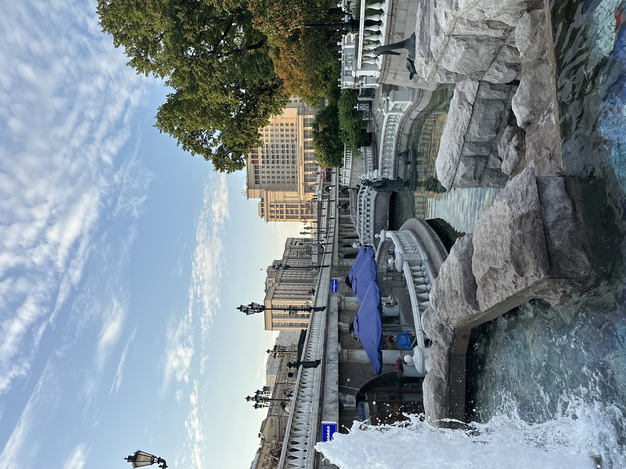 The image depicts a view of the Alexander Garden in Moscow, Russia, facing the historic Hotel Moskva. In the foreground, a large fountain splashes water into a clear pool, with rough-hewn rocks lining the edges. Beyond the fountain, a tiered stone structure features white balustrades and decorative lamp posts. Several blue umbrellas are set up over tables and chairs, and a woman in a blue shirt is seated near them.
Further back, a bridge spans a section of the garden, and a woman is seen walking across it. In the background, the imposing facade of the Hotel Moskva, a neoclassical building, dominates the skyline. Green trees and other buildings are visible, along with a bright blue sky filled with scattered clouds. The scene captures a tranquil outdoor atmosphere in a prominent Moscow location.