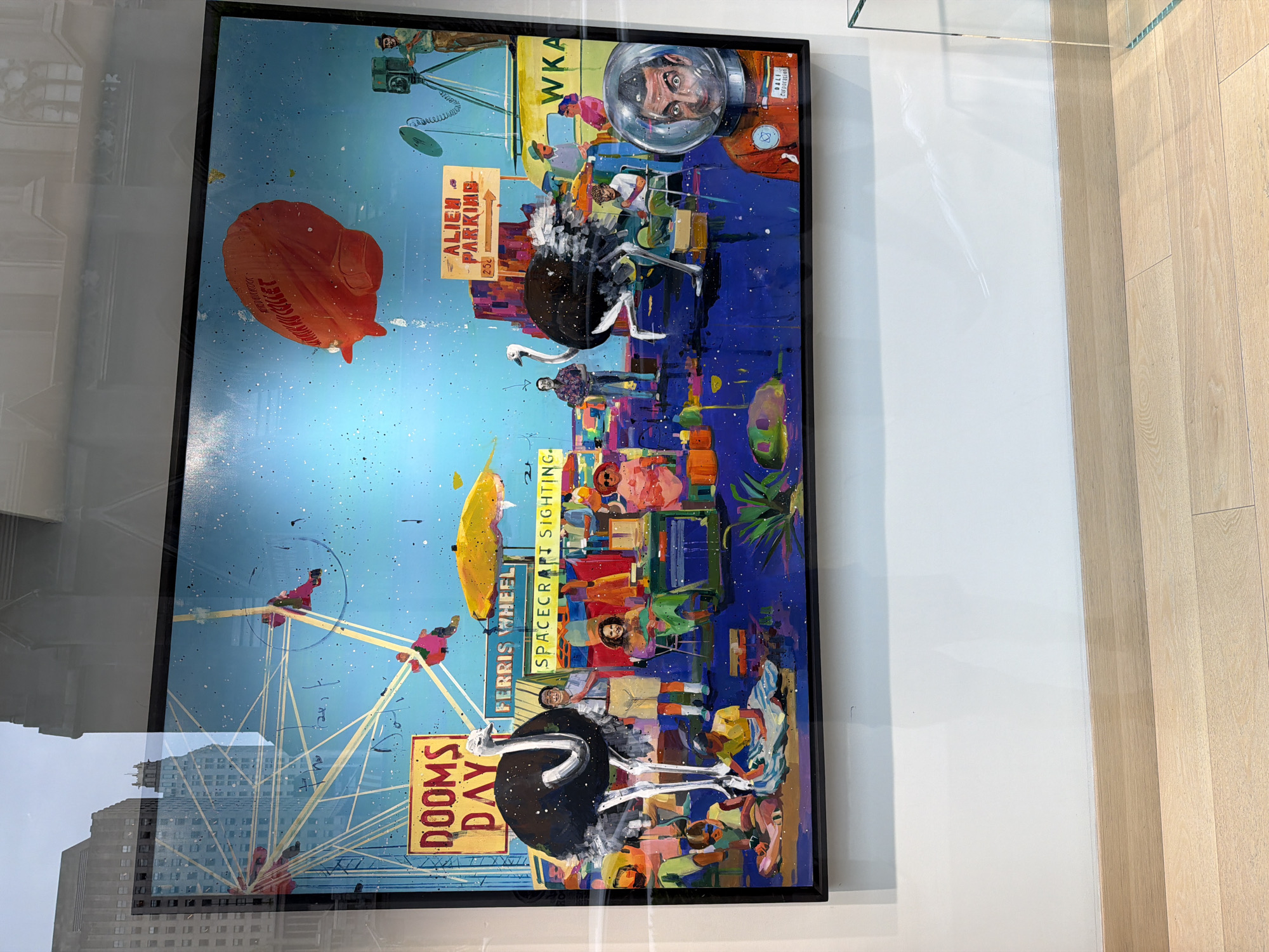 The image shows a framed art piece displayed in what appears to be a storefront window, possibly in New York City. The art itself is a whimsical, surreal composition. It features a Ferris wheel with figures riding in the carts, ostriches, and various signs like "Dooms Day," "Spacecraft Sighting," and "Alien Parking." Other elements include a blimp or airship and a person in an astronaut helmet. The city's skyline is reflected in the window, suggesting an urban location. The environment includes a white wall behind the framed artwork and a light-colored wooden floor visible beside the wall.