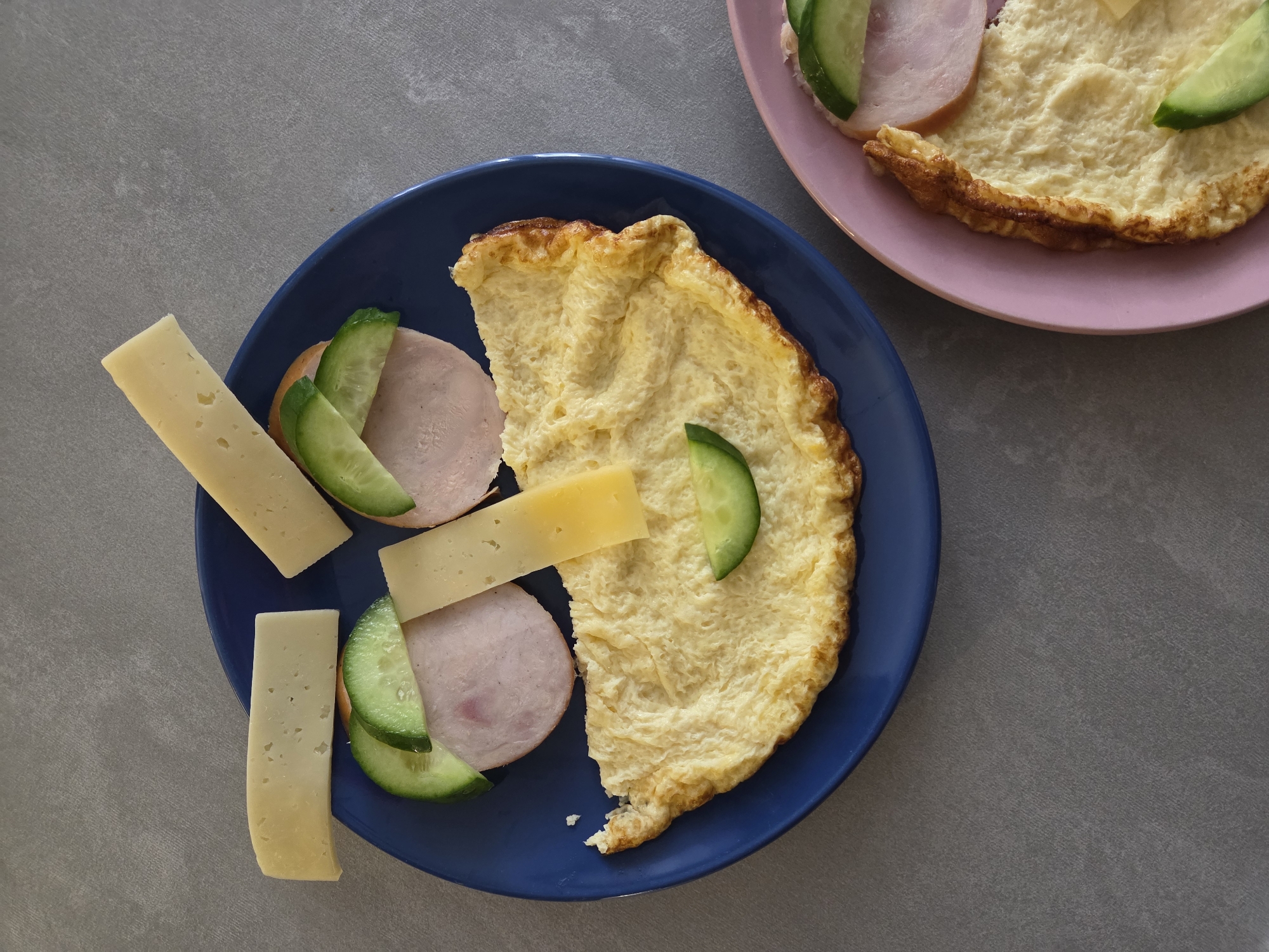 An overhead view captures two plates of a prepared meal, likely breakfast or brunch, set against a gray, textured surface in what is presumed to be Birobidzhan, Russia.
In the foreground, a round, deep blue plate holds a substantial serving. A large, fluffy, golden-yellow omelet, characterized by its textured surface and slightly browned, crispy edges, dominates the center. Accompanying the omelet are several components: multiple rectangular slices of pale yellow cheese with small, irregular holes; thin, round slices of light pink ham or deli meat; and vibrant green cucumber slices cut into wedges. These items are neatly arranged around the omelet.
Partially visible in the upper right corner is a portion of a second, round, pink plate. This plate also features a section of a similar golden omelet, topped with a round slice of ham and a green cucumber wedge, indicating a consistent meal offering.
The background is a uniform medium-gray, showing a subtle, fine texture, possibly resembling concrete or a laminate countertop. The lighting is bright and even, suggesting a daytime setting. No individuals are present in the image, and there is no visible text. The arrangement of the food suggests a simple, hearty, and appealing meal.