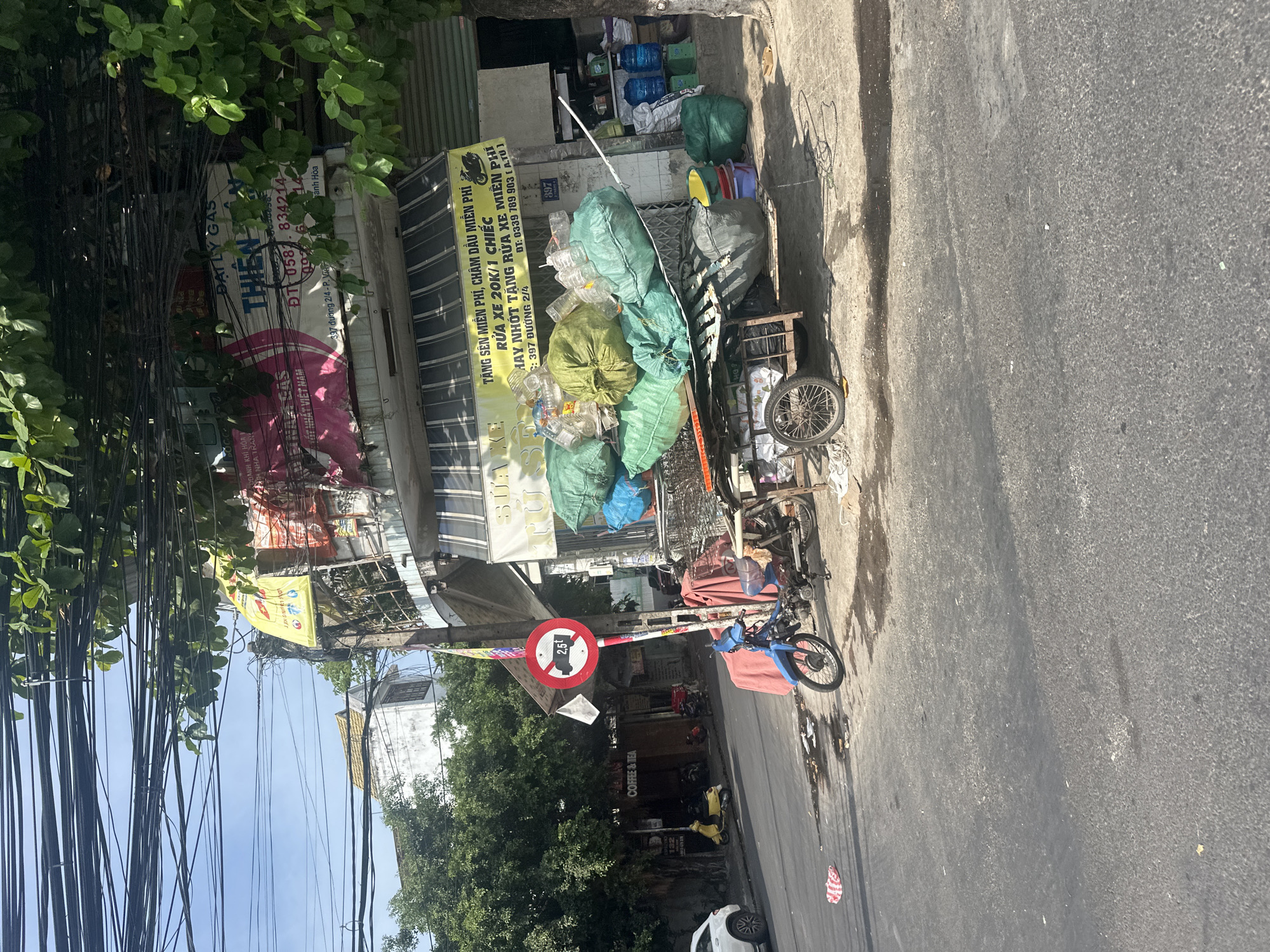 The image depicts a street scene in Bac Nha Trang Ward, Vietnam. A street vendor is seen pushing a cart loaded with green and blue plastic bags, likely filled with recycled materials, on a paved road. A blue motorcycle is parked next to the cart, and a person is seated on it, wearing a dark-colored headscarf and shirt. In the background, a multi-story building with various signs is visible, including one advertising "THIEN THAN" and another for "DAILY GAS." A "2.5t" weight limit sign is posted on a pole. The area appears to be a commercial or mixed-use zone with overhead electrical wires, trees, and other vehicles present. The overall environment suggests an urban setting with everyday activity.