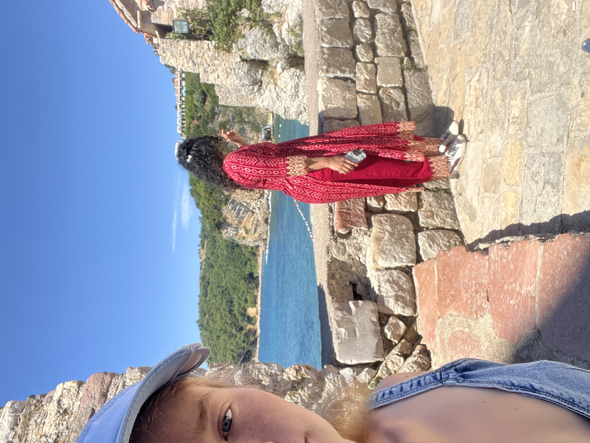 This is a medium shot, taken from a slightly low angle, of two women outdoors in Budva, Montenegro. The woman on the left, facing the camera, is only partially visible from the chest up. She is wearing a light blue baseball cap and denim overalls. Her left eye and a portion of her nose and cheek are visible. The woman on the right, facing away from the camera and looking out at the sea, is wearing a vibrant red, patterned tunic over red pants. She has dark, curly hair and is holding a smartphone in her right hand.
They are standing on a stone pathway, with a low stone wall separating them from a body of blue water. Beyond the water, a green, tree-covered hillside with buildings can be seen, and the clear blue sky stretches above. The architecture of the buildings in the background suggests a coastal, possibly historic, location. The rough-hewn stone of the pathway and wall, along with the natural landscape and sea, indicate an outdoor setting within the historic old town or along the coast of Budva. The sunlight suggests it is daytime, and the bright blue sky and clear water indicate fair weather. The scene appears to be a tourist destination, given the presence of people enjoying the view.