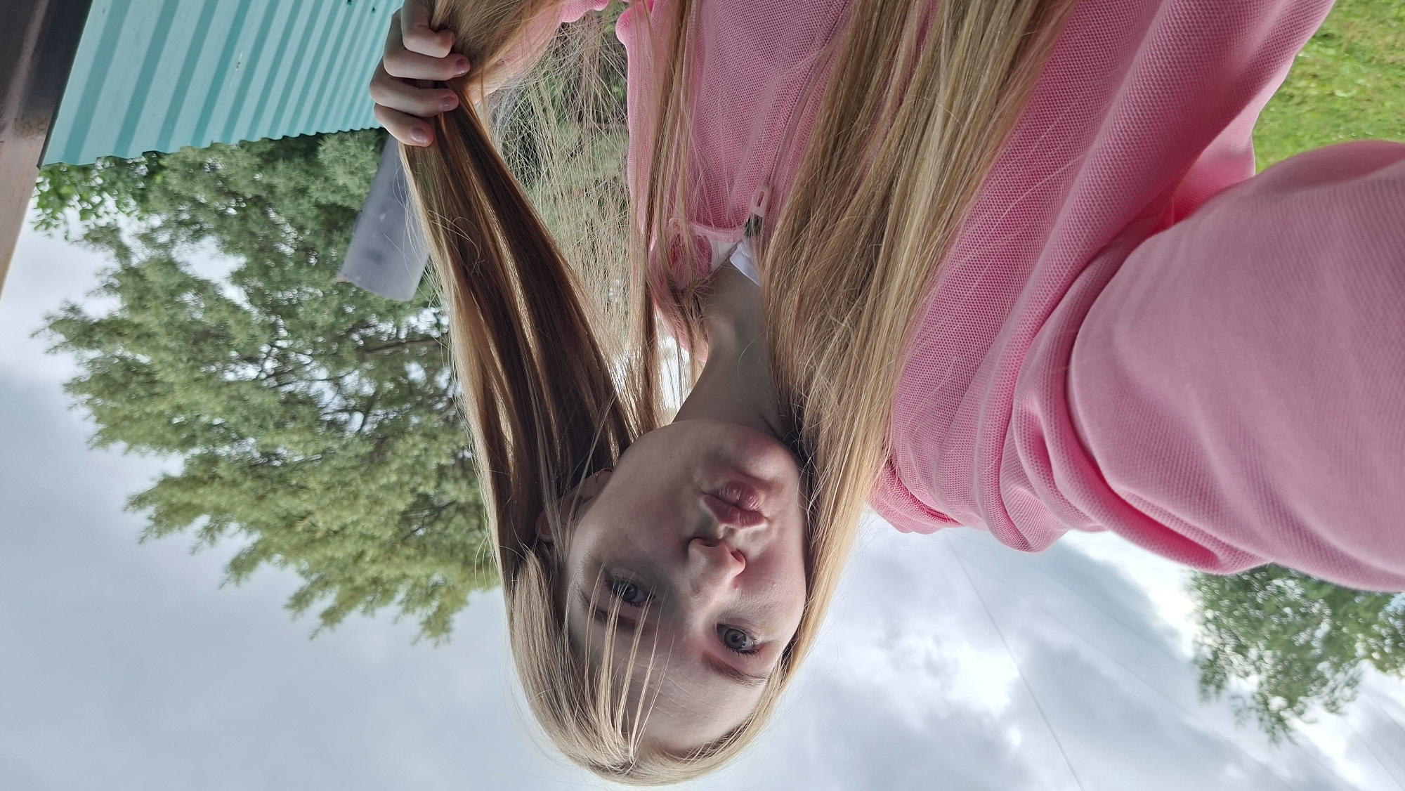 A young girl with long blonde hair, wearing a pink polo shirt, is captured in a selfie. Her face is turned towards the camera, with her lips puckered in a kiss pose. She is holding a section of her hair with her left hand. The background suggests an outdoor setting with lush green trees and a cloudy sky. A glimpse of a turquoise corrugated metal structure is visible on the left edge of the frame. The lighting indicates it's daytime. The overall mood appears playful and lighthearted. The image was taken in Bolshoy Bukor, Russia.