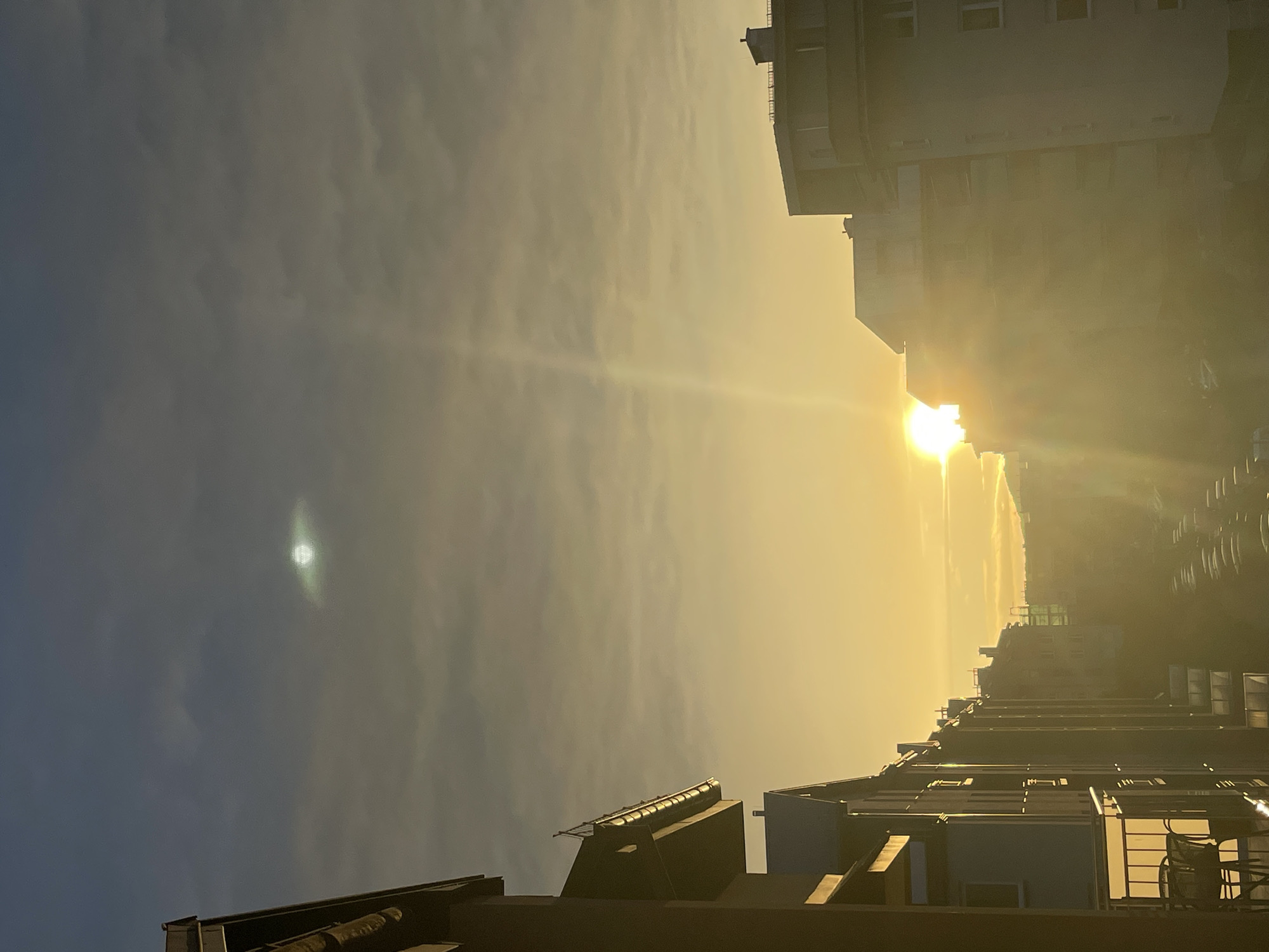 The image captures an urban landscape at either sunrise or sunset, viewed from an elevated position, likely a balcony or window. The sky is largely overcast with a mix of grey-blue and warm golden hues, indicative of a low sun angle. In the upper-center sky, a distinct, circular, bright green luminous orb with a soft halo is visible. The sun, a brilliant white-yellow disk, is positioned low on the horizon, partially obscured by buildings on the right side of the frame, emitting strong golden light rays that create prominent lens flares and atmospheric haze across the lower-right and center.
On the left, a multi-story residential building with a dark, reddish-brown facade and multiple balconies with dark railings dominates the foreground. Its roofline and gutter system are visible at the top left. A row of similar buildings recedes into the distance along what appears to be a street. On the right, several light-colored, multi-story buildings are heavily backlit and partially silhouetted by the intense sunlight, their windows and roof structures faintly discernible.
Between the rows of buildings, a street extends into the background, where numerous parked cars are visible as indistinct shapes due to the strong light and distance. Distant hills or mountains with some vegetation are faintly visible on the far horizon, beyond the urban structures. The overall scene is devoid of discernible people. The dominant lighting is warm, golden, and hazy, transitioning to cooler tones in the upper sky, with a notable vertical light shaft extending upwards from the sun on the right.