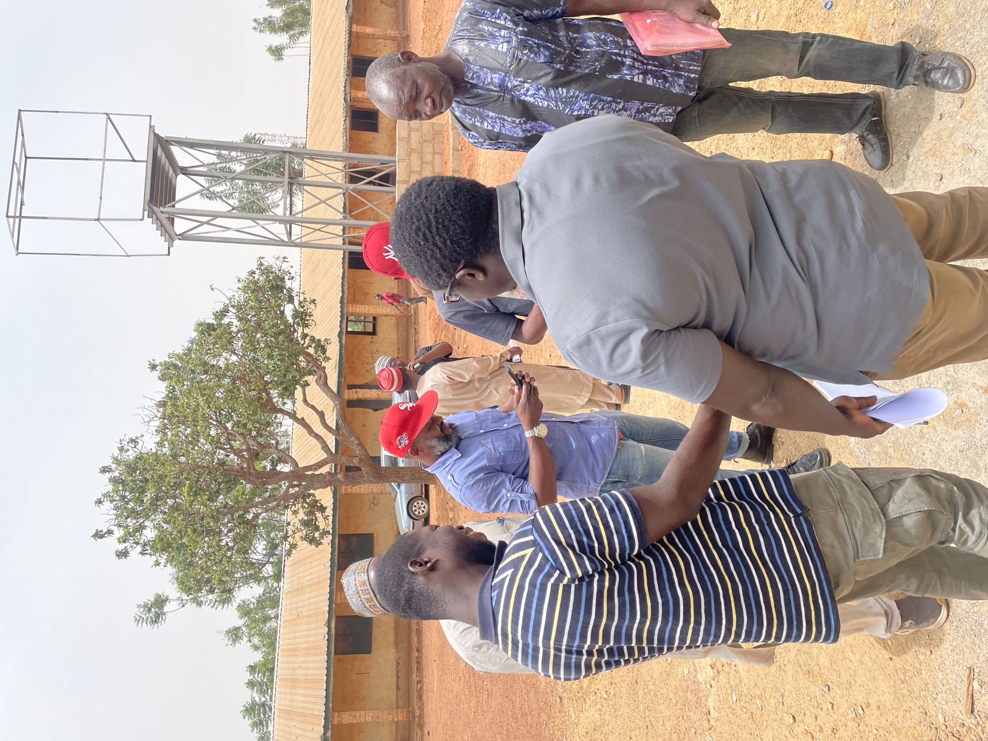 A group of men are gathered outdoors in Jos, Nigeria. The foreground depicts a man wearing a striped blue and yellow shirt with green pants. Behind him, a man in a blue shirt and red cap holds a phone. A tree and a water tower structure are visible in the background, with a building behind them. The ground appears to be dirt and gravel.