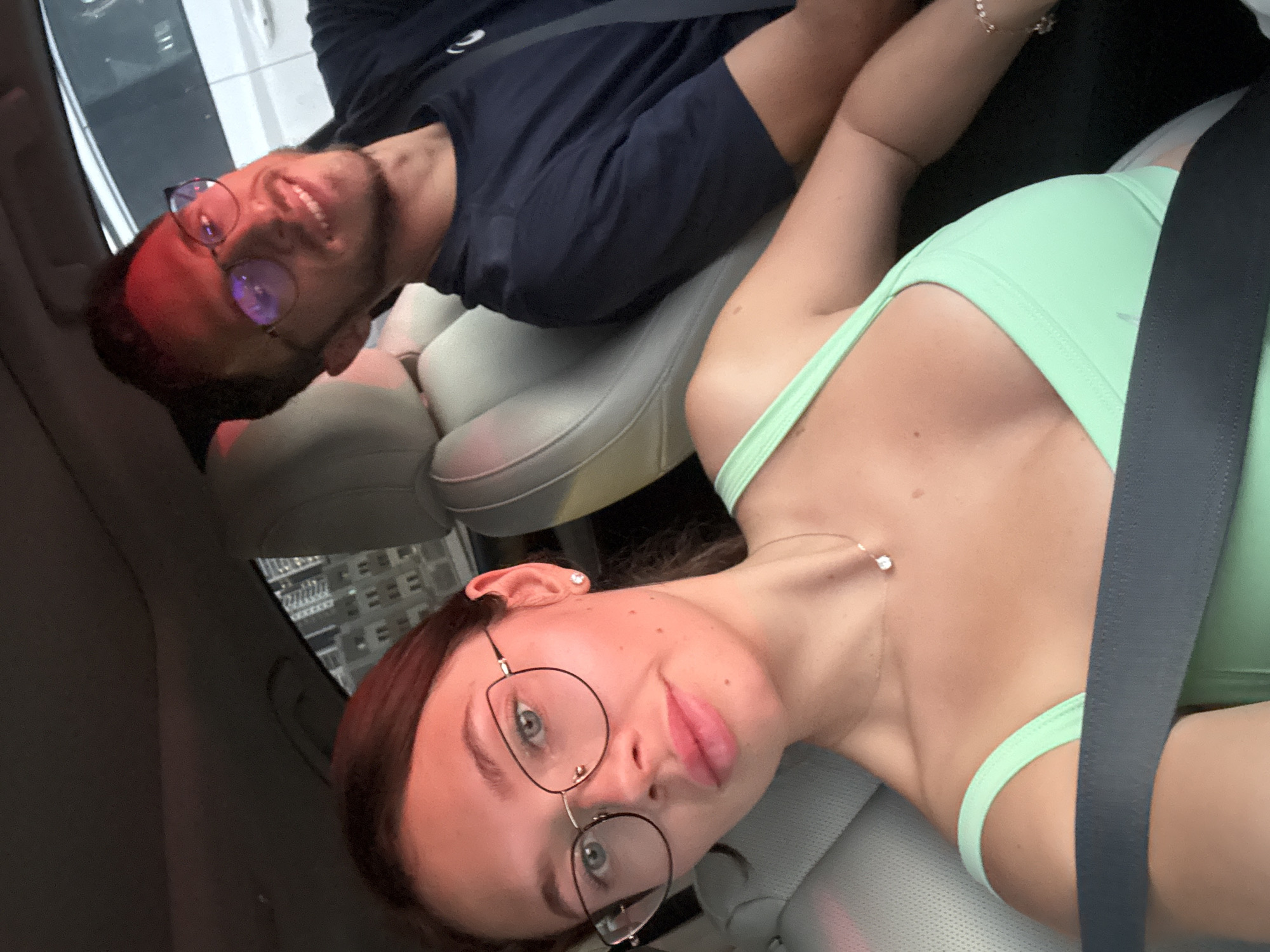 A man and a woman are in a car, both wearing seatbelts. The man is wearing glasses and a dark blue shirt. The woman is wearing glasses and a light green top. The building outside of the car indicates they are in Tel Aviv, Israel.