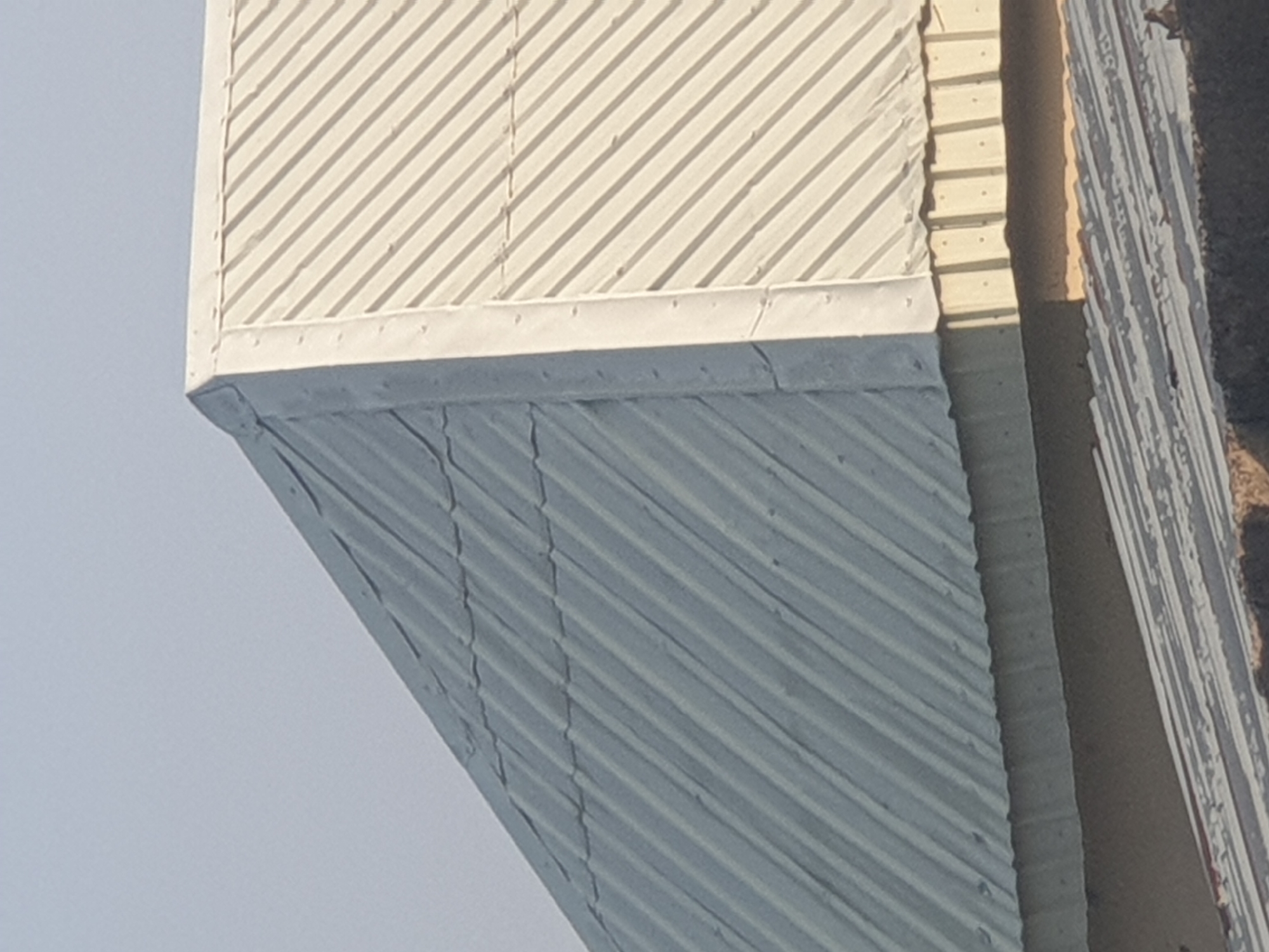 The image shows a close-up view of two adjacent corrugated metal roofs under a clear, pale blue sky. The roof in the foreground, on the left side of the image, is a dull gray color. It appears older and shows some signs of wear, with visible seams and minor undulations. The roof on the right side is a lighter, off-white color and has a more uniform, cleaner appearance. This roof has a distinct ridge where the two sections meet, which is capped with a white metal flashing secured with visible rivets. The angled ridges of the corrugated metal run diagonally from the bottom left to the top right of both roofs. The lower part of the image reveals the edge of a lower structure, also with a corrugated roof, but the details are less clear. The overall scene suggests an urban or suburban environment with a focus on architectural details of roofing materials.