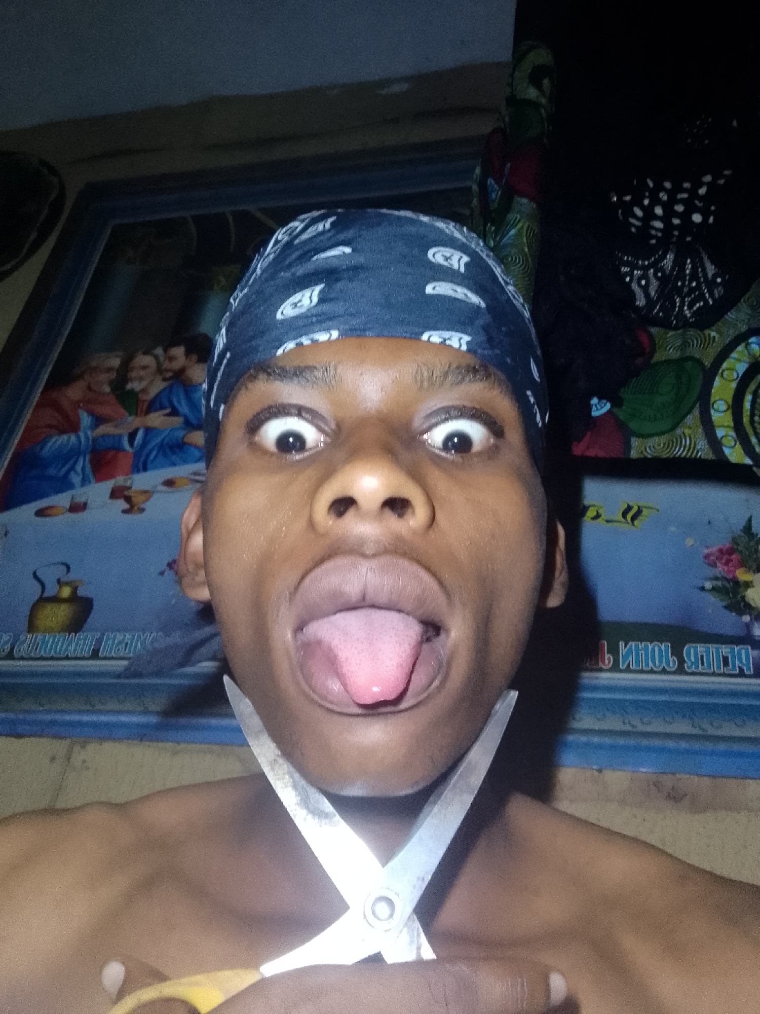 A person wearing a dark-colored bandana is holding scissors under their chin and sticking out their tongue. Behind the person is a framed picture of the Last Supper and other objects out of focus. The location is Unknown, Unknown.
