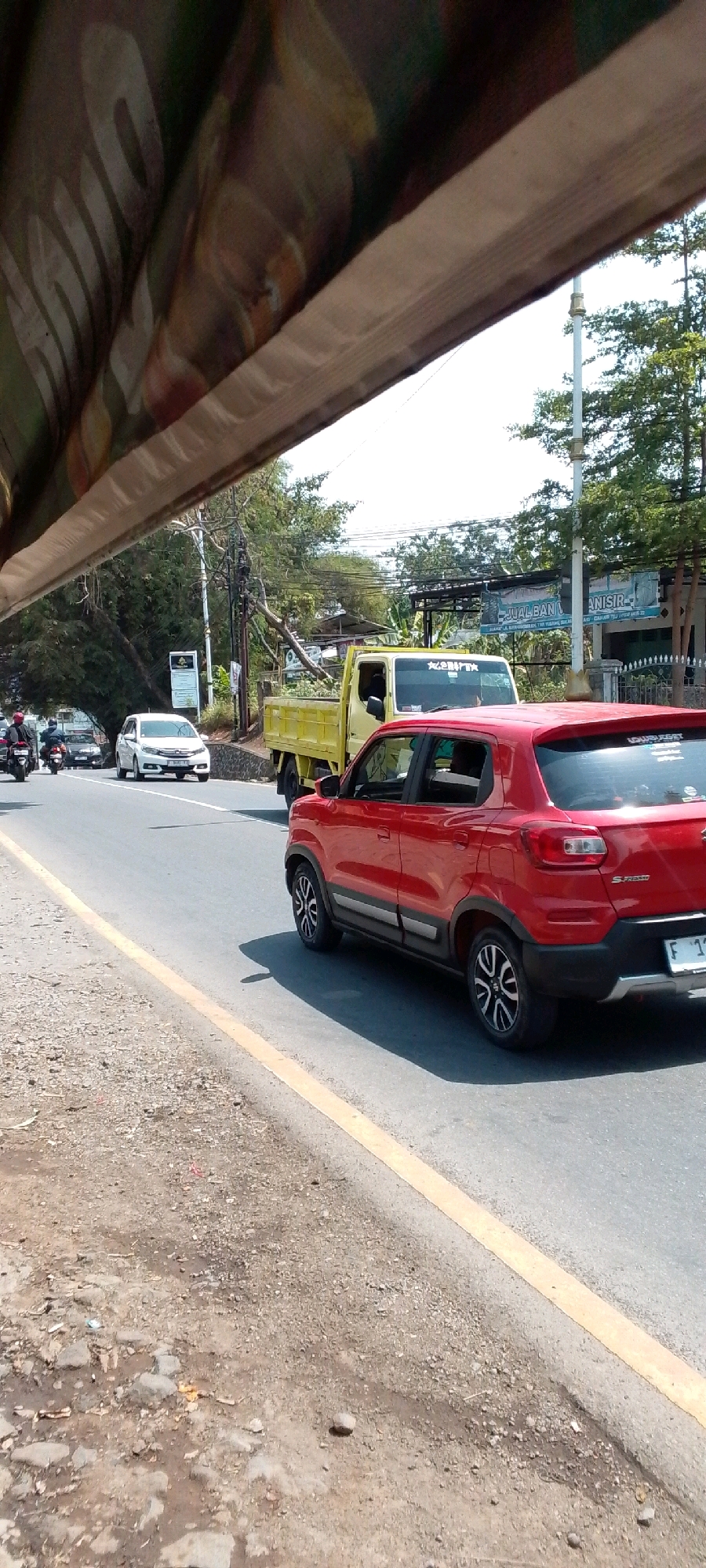 The image captures a street scene in Cilaku, Indonesia. In the foreground, a red car is parked on the right side of the road, with its rear visible. The road surface appears to be asphalt with a yellow line marking the edge. To the left of the road, there is a rough, gravelly shoulder.
Further down the road, a yellow truck is driving away from the camera. In front of the truck, a white car is also in motion. Behind the white car, two motorcycles are visible, with riders wearing helmets.
The background shows trees, utility poles, and some buildings. The sky is bright and appears to be clear, suggesting a sunny day. A canopy or awning is present in the very top of the image, obscuring a portion of the sky and trees. Some signage is visible on the buildings in the distance, including what appears to be a banner with Indonesian text. The overall impression is of a typical urban or semi-urban street with moderate traffic.