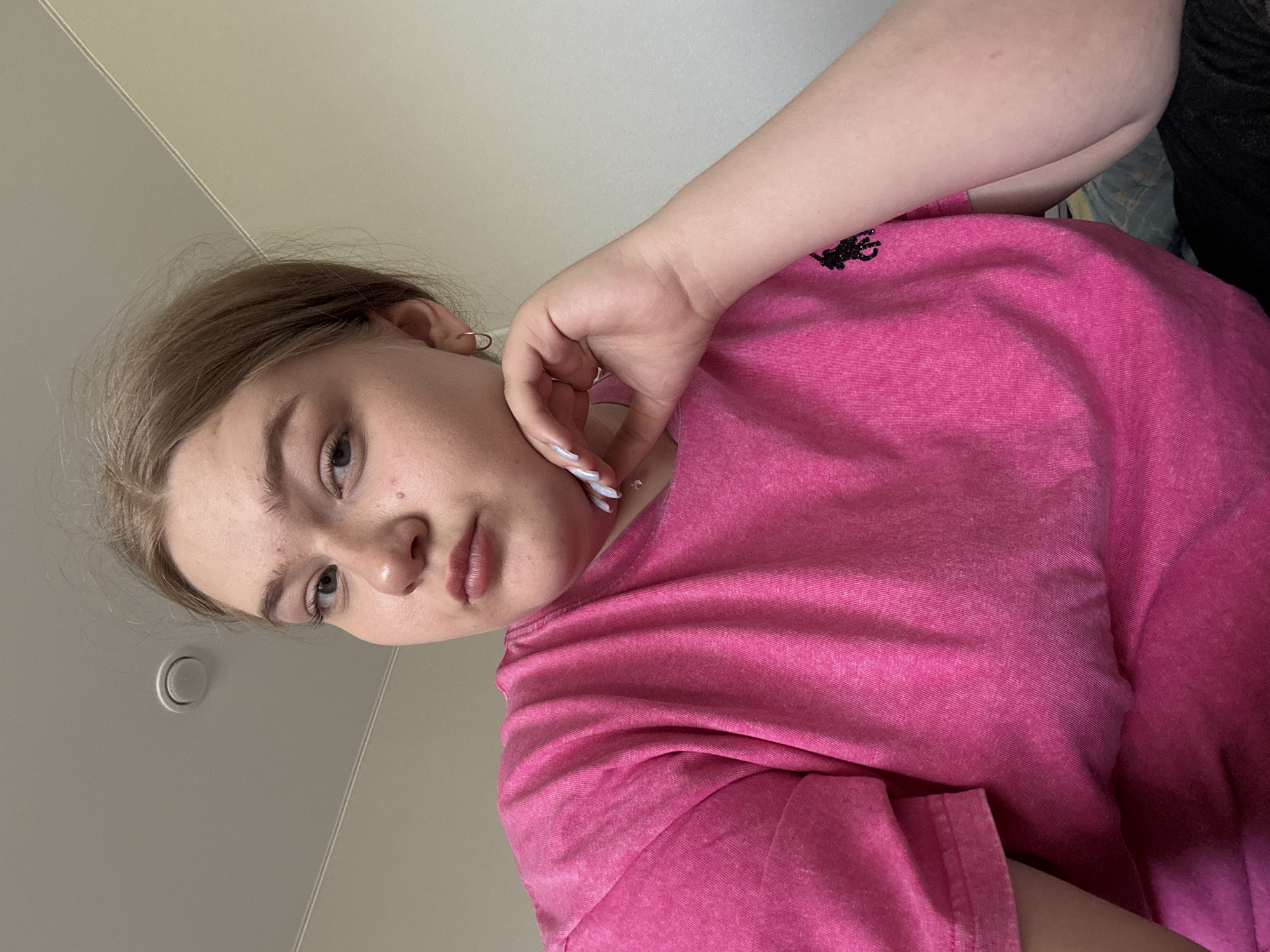 A young woman is angled towards the camera. She is wearing a pink t-shirt with a black logo. Her right hand is raised to her face. The background is a neutral beige wall with a light fixture. The location is unknown and there is no evidence that this image was taken in Фоки, Russia.