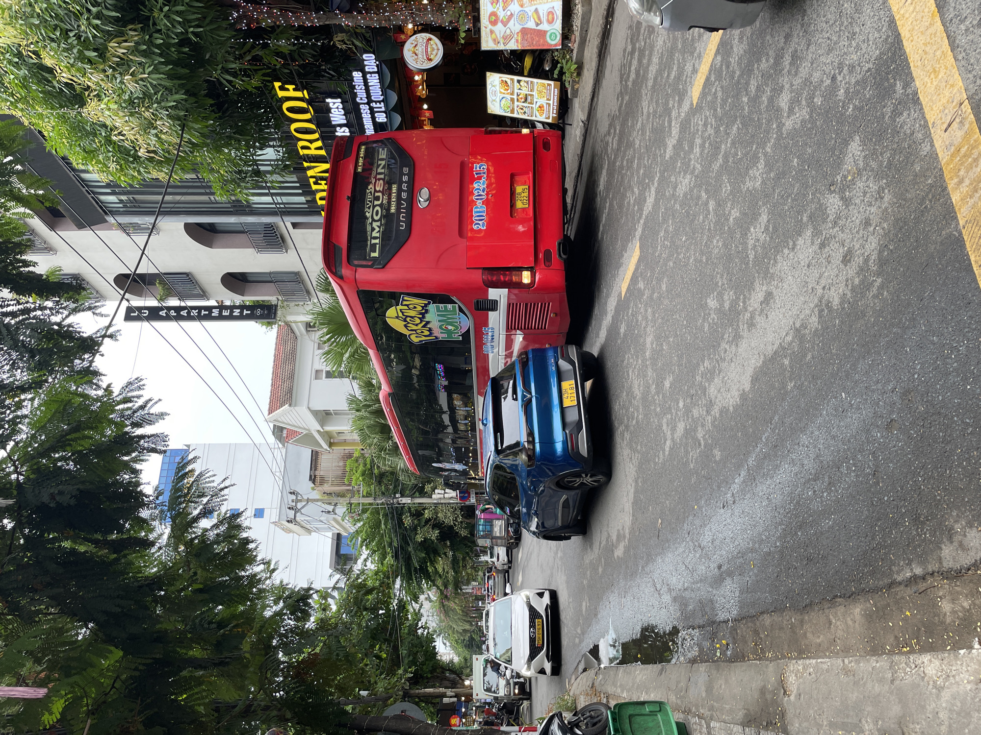 The image depicts a street scene in Da Nang, Vietnam, likely on a cloudy day. The foreground features a wet asphalt road with yellow lane markings. On the right side of the road, a blue SUV is parked, and a portion of a silver car is visible. A large red bus, advertising "Pokemon HOME" and "LIMOUSINE UNIVERSE," is positioned in the middle of the road, partially obscuring the view of other vehicles behind it. The bus has a license plate that reads "20B-022.15". To the right of the bus, a building with a sign for "GOLDEN ROOF" is visible, with illuminated decorations and menus displayed outside. Further down the street, more vehicles, including a white sedan with license plate "75H-016.93", a white van, and a truck, are seen in traffic. Trees with dense green foliage line the left side of the street, and buildings of varying architectural styles are present in the background, including one with the prominent signage "LU APARTMENT". A green trash bin is located on the left sidewalk. The overall scene suggests a typical urban environment with active traffic.