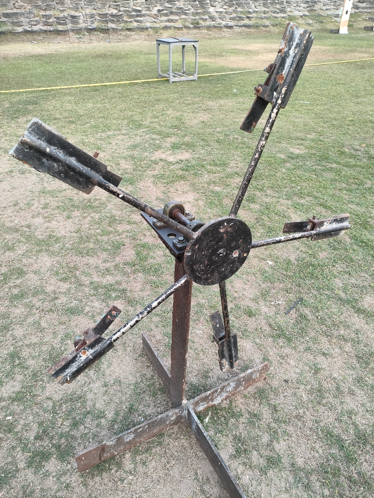 A rusted, four-bladed metal contraption stands on a grassy field in Banguntapan, Indonesia. The contraption consists of a central circular disk from which four arms extend outwards, each ending in a bladed element. The entire structure is mounted on a metal base. In the background, a grassy area is bordered by a raised, tiered structure, possibly a stadium or amphitheater, made of stone or compacted earth. A yellow line runs across the grassy field in the middle ground, and a small metal table with a stool is visible further back. The overall scene suggests an outdoor recreational or training area.