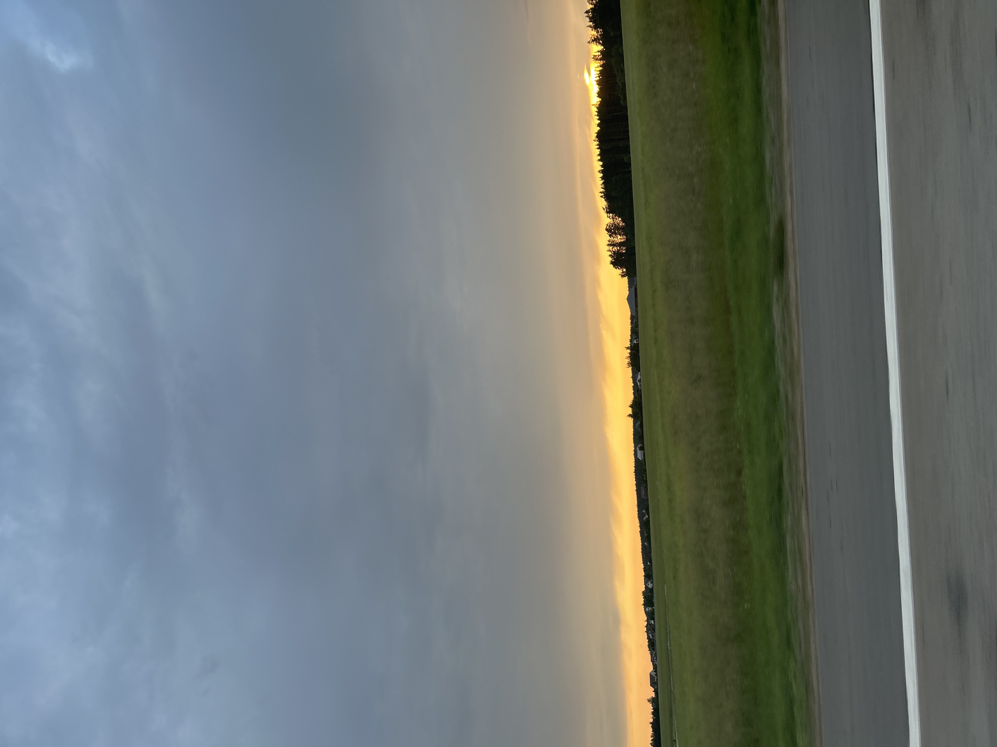 The image depicts a landscape scene at dusk. The sky is filled with a mix of dark, cloudy grey at the top, transitioning to a bright orange-yellow glow near the horizon, suggesting sunset or sunrise. Below the sky, a wide, green field stretches across the middle ground, with hints of darker green suggesting variations in the grass or perhaps shadows. In the distance, along the horizon line, a dark silhouette of a coniferous forest runs from left to right. Scattered within the field and along the edge of the forest are several small, light-colored buildings, indicative of a rural settlement. The foreground of the image is dominated by a grey asphalt road, with a distinct white line running along its edge. The road and the field appear slightly blurred, suggesting the photo was taken while in motion, likely from a vehicle. The specific location is identified as the Ozyaritska-Slabadski rural council in Belarus. There are no people, animals, or specific actions visible in the scene. The environment is natural with man-made elements in the form of buildings and a road.