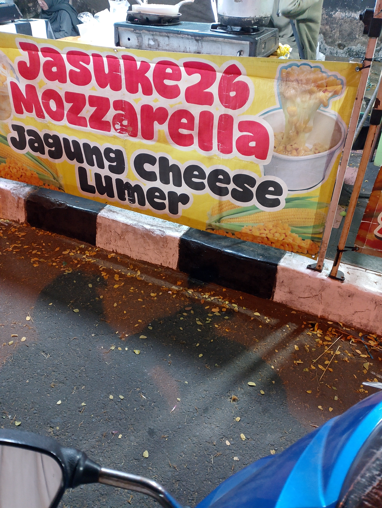 This is a close-up, eye-level shot of a food stall banner, likely in Magelang, Indonesia, given the signage. The banner is yellow and features bold red and black text. The prominent red text reads "Jasuke Mozzarella" and "26". Below this, in black text, it says "Jagung Cheese Lumer". A graphic of melted cheese oozing from a bowl of corn kernels is also visible on the right side of the banner. The banner is attached to a wooden pole. In the foreground, a portion of a black and white striped curb is visible, with fallen leaves scattered on the dark, wet asphalt below. A reflection of what appears to be a motorcycle handlebar and a blue motorcycle fairing is present in the bottom left and right corners, indicating the photograph was likely taken from the perspective of someone on a motorcycle. The background is blurred but suggests a street scene with a person in a headscarf in the upper left and a food preparation area with a stove to the right of the banner.