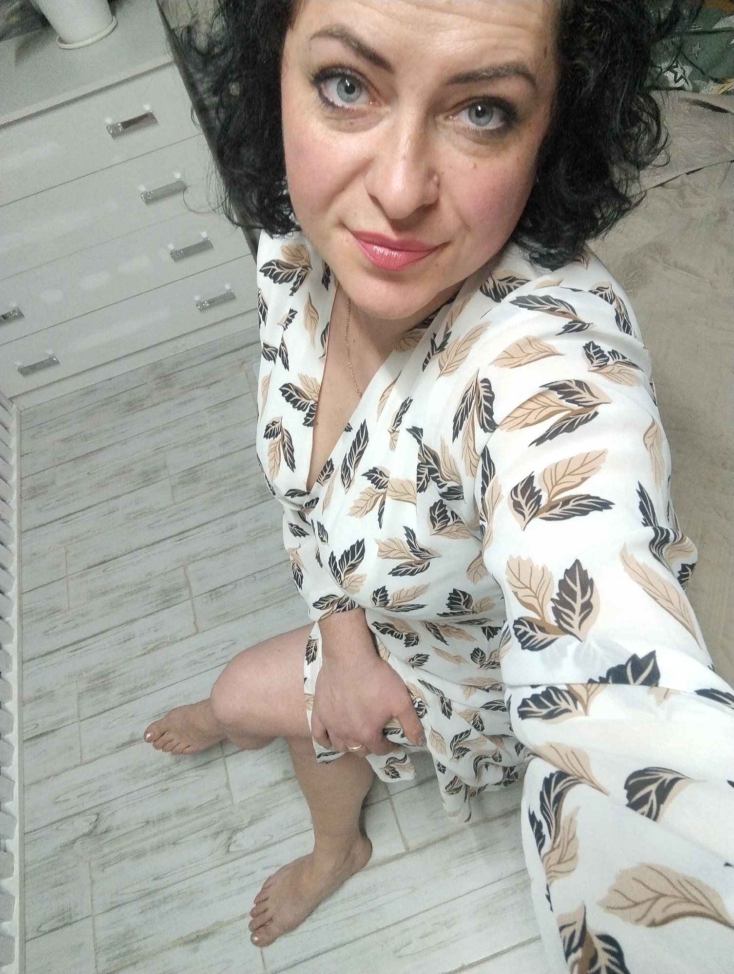 A woman with dark, curly hair and striking blue eyes is captured in a high-angle, close-up photograph taken indoors. She looks directly at the camera with a calm, slightly smiling expression, her lips painted with light pink lipstick. Her fair complexion shows a touch of blush on her cheeks.
She is wearing a loose-fitting, light-colored garment, possibly a dress or a long top, featuring a repeating pattern of stylized leaves in shades of brown, tan, and dark grey or black. A thin, delicate necklace is visible around her neck, and a simple ring adorns a finger on her right hand, which rests casually on her lap. She is barefoot, with light-colored polish on her toenails.
The woman is seated or kneeling on a floor covered with light-colored, wood-effect tiles or laminate flooring. In the background, to her left, a white chest of drawers or cabinet with multiple silver handles is visible. A white pipe or fixture can be seen partially above the cabinet. The setting appears to be a domestic interior in Grishanki, Russia. No visible text is present.