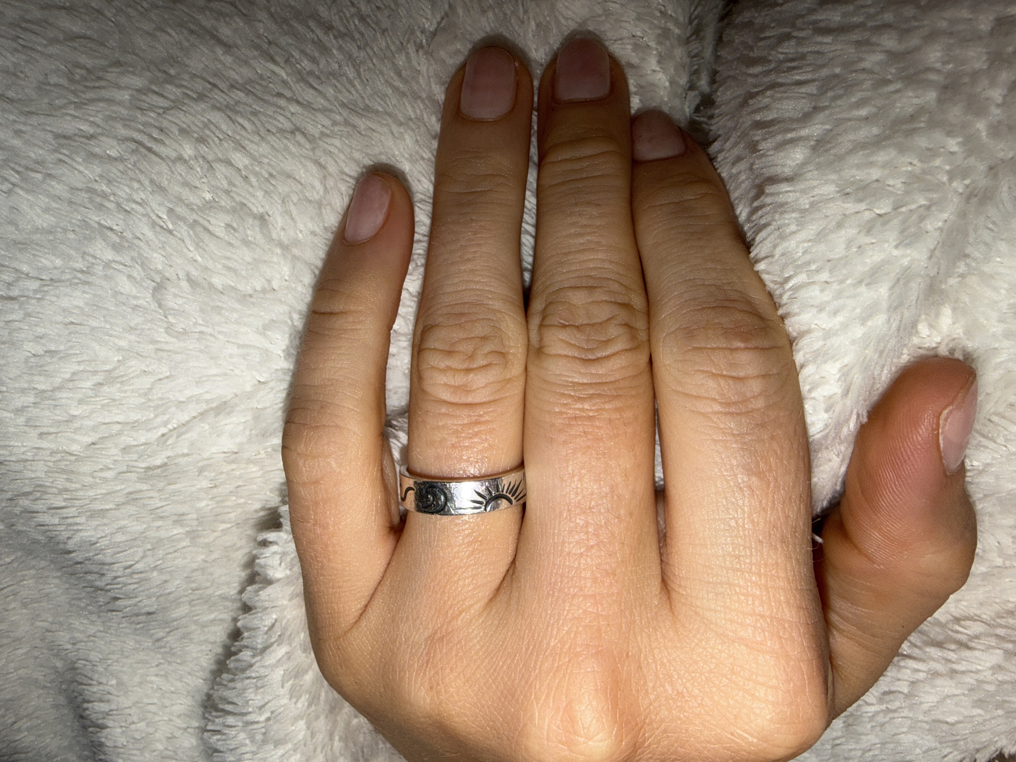 A hand is resting on a white, fluffy surface. The hand has a silver ring with sun and moon designs on the ring finger. The fingernails are unpainted. The image does not include location information and therefore it is impossible to provide location information.