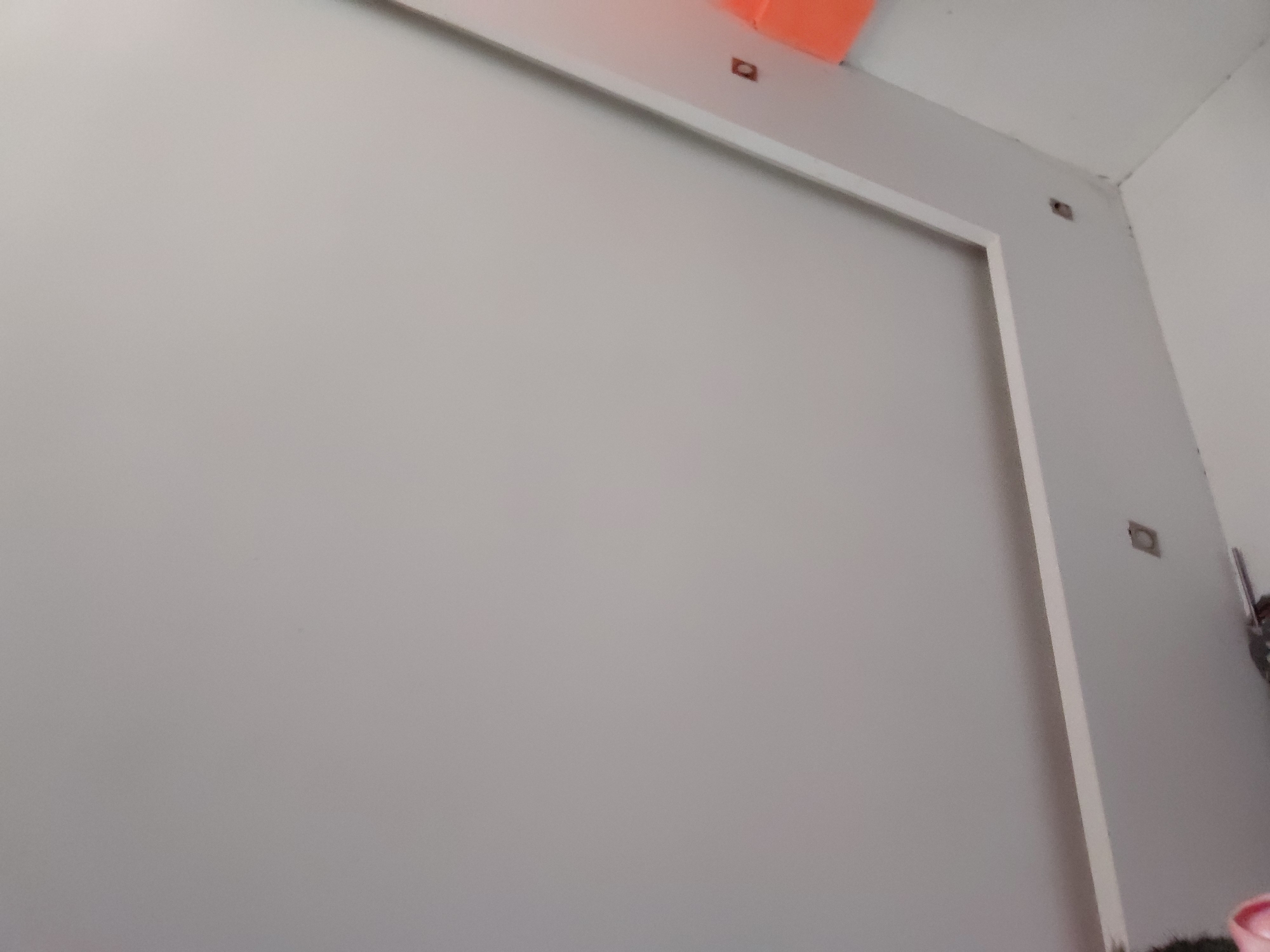 The image displays an interior view of a room, focusing on a large, light grey wall. A prominent white trim outlines a rectangular section of this wall, suggesting a decorative panel, a built-in feature, or a designated space for an object like a television. In the upper part of the image, an orange object is partially visible, possibly a light fixture or a structural element, positioned near the ceiling line.
Several small, dark, rectangular cutouts or recesses are visible on the white trim and the adjacent wall in the upper right corner, likely intended for electrical outlets, switches, or mounting hardware. The corner of the room is visible on the right, where two light-colored walls meet the ceiling, all appearing to be painted in shades of white or very light grey.
In the bottom right corner, a portion of a pink container, possibly a bucket, is visible, along with some dark, textured material that might be artificial grass or a similar decorative element. Another indistinct object is partially visible further to the right. The room appears clean and minimalist, possibly indicating a recently finished construction, a space awaiting decoration, or a contemporary design choice. No people are present, and no activity is occurring. There is no visible text. The lighting is uniform, typical of an indoor environment without direct sunlight.