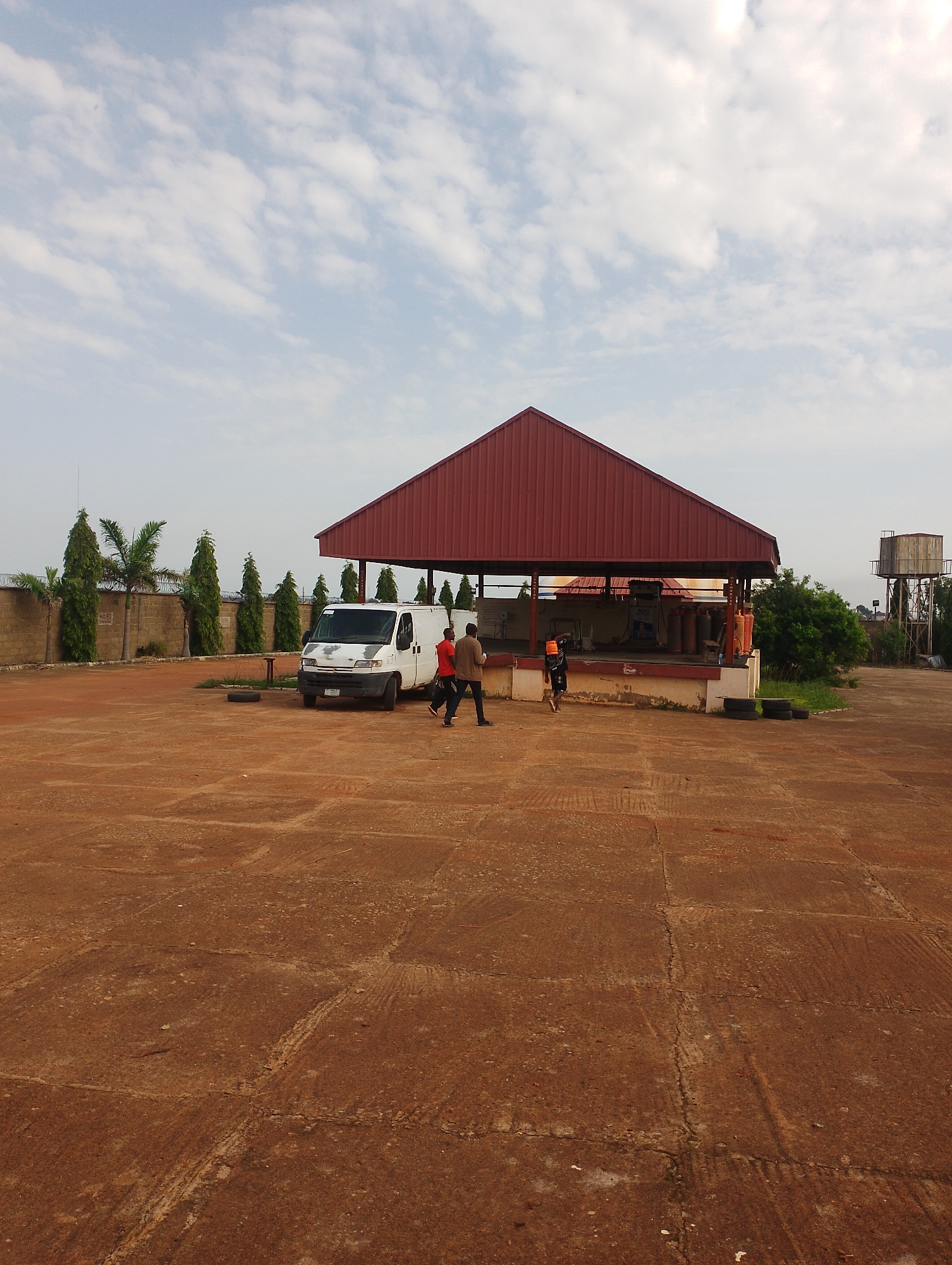 The image depicts an outdoor industrial area with a large, open-air structure covered by a red, gabled roof. In front of this structure, a white van is parked. Two men are standing near the van, one wearing a red shirt and the other a brown shirt, appearing to interact or walk. To the right of the structure, there is dense green foliage and a tall water tower in the distance. The ground is a reddish-brown, paved surface with visible cracks and tire marks. A low stone wall runs along the background on the left, topped with barbed wire, and several tall, conical trees are planted behind it, along with some palm trees. The sky is partly cloudy, with white clouds against a light blue backdrop.