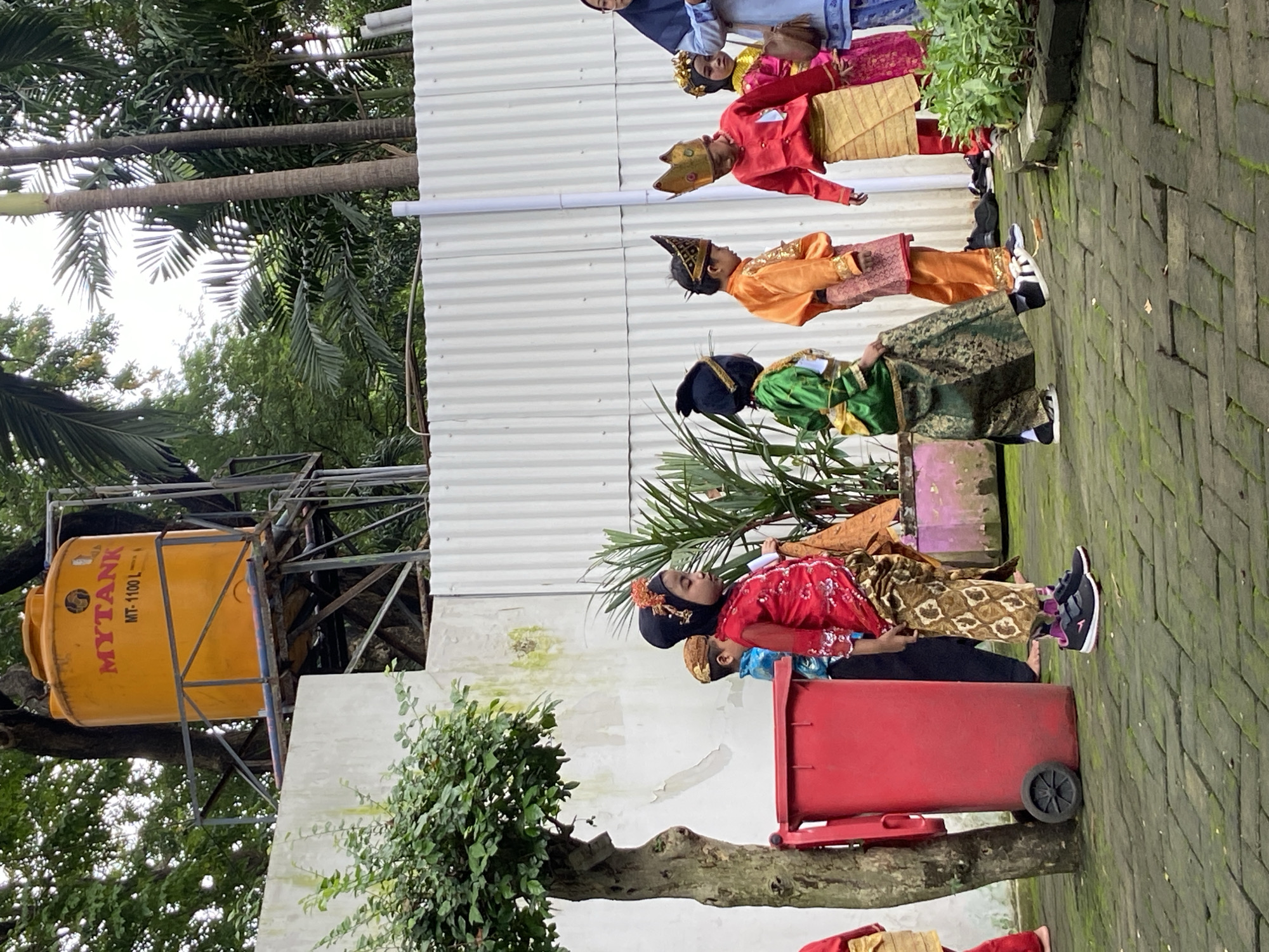 This outdoor image, taken in Surabaya, Indonesia, features a group of children and at least one adult adorned in vibrant traditional Indonesian costumes. The scene unfolds in what appears to be a paved courtyard or schoolyard, with a white corrugated metal wall serving as a backdrop. Lush green tropical foliage, including prominent palm trees and other plants, frames the top and left sides of the image. The brick pavement is visibly mossy, suggesting a humid environment.
The children are dressed in elaborate ceremonial attire, showcasing a variety of colors including red, gold, orange, and green, complemented by distinct headdresses and patterned sarongs. From left to right, visible children include one in a red and gold outfit with a golden crown-like headdress, another in an orange and gold costume with a tall, pointed black and gold headdress, and a third in a green and gold outfit with a black cap. Further right, a child in a red top and patterned sarong, with a gold headband, is seen near a red wheeled trash bin. Modern athletic shoes worn by some children provide a contemporary contrast to their traditional garments. An adult wearing a light blue hijab and accompanying another child in a pink costume is partially visible in the background.
The children appear to be gathered for a cultural event, performance, or procession, given their ceremonial dress. They are lined up along the wall, possibly awaiting their turn or posing. The lighting suggests it is daytime, with soft light indicating an overcast sky or early morning/late afternoon. On the far left, a large yellow water tank on a metal stand is visible amidst the trees, bearing the text "MYTANK MT-1100 L". The overall impression is one of cultural display and anticipation.