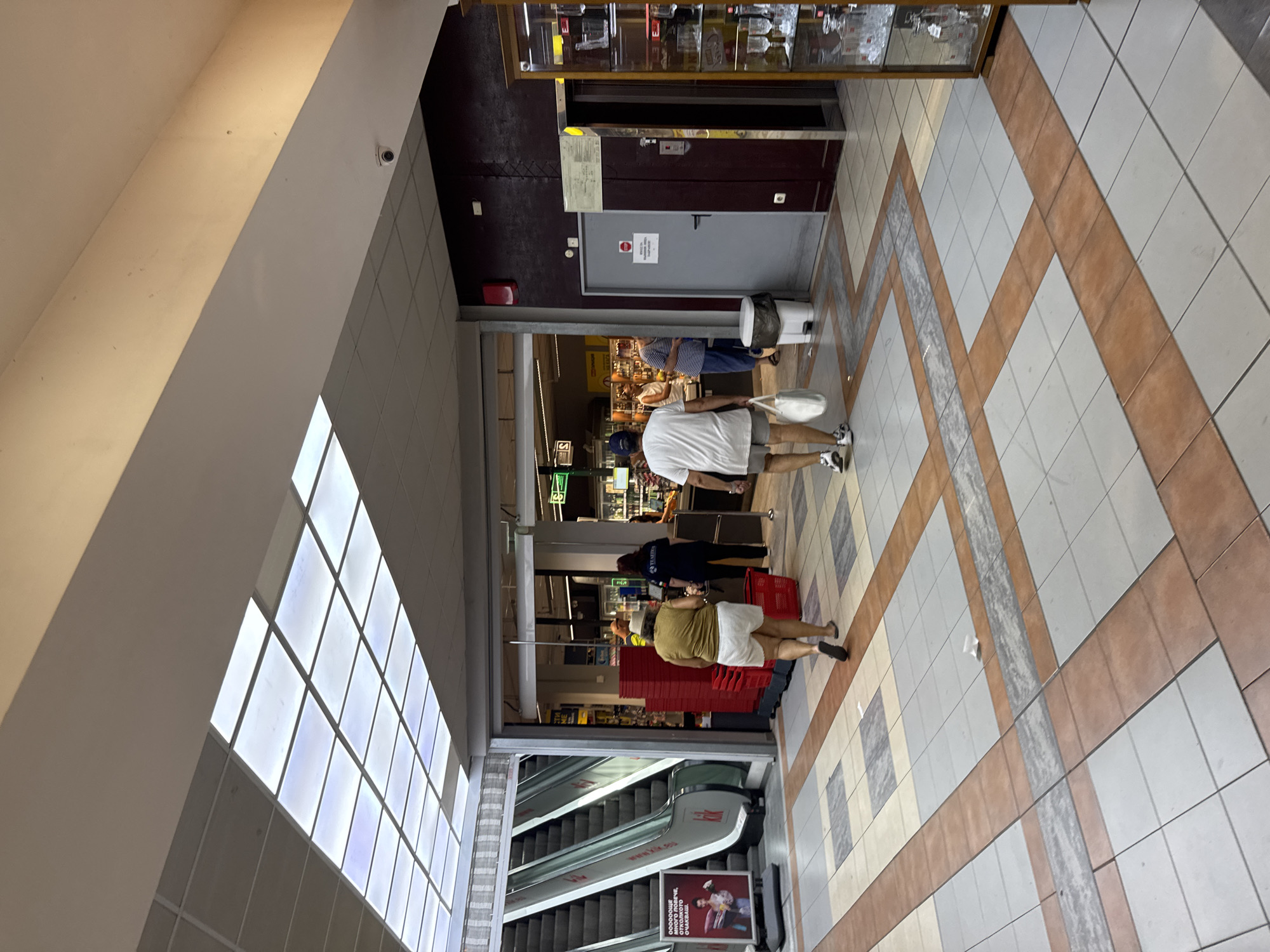 The image depicts the interior of a shopping mall or supermarket in Burgas, Bulgaria. In the foreground, a tiled floor with geometric patterns is visible. Two escalators are on the left, with a red and white advertising poster next to them. Several people are walking through the mall. One woman in a yellow top and white shorts is pushing a red shopping basket. A man in a white t-shirt and grey shorts walks away from the camera carrying a white bag. Other shoppers and employees are visible in the background, some near checkout counters. The ceiling features a grid of fluorescent lights. A door and an elevator are visible on the right side of the image, along with a display cabinet filled with glassware. The overall environment appears to be a public indoor commercial space.
