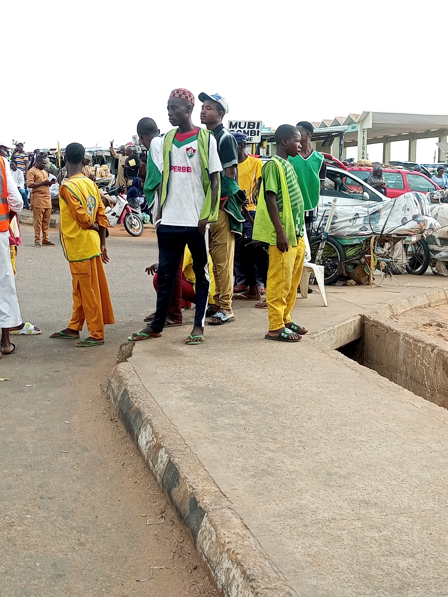 In Gombe, Nigeria, a group of young men stand on a paved area next to a drainage ditch. One man in the foreground wears a white t-shirt with "PERBEI" printed on it and a green vest, a patterned red and white kufi on his head. To his right, another young man in a green vest and yellow pants looks away. Further left, a person in a yellow tunic and orange pants stands with their back to the camera. Other individuals are visible in the background, some near a motorcycle and several vehicles, including a red car. The environment appears to be an outdoor market or transportation hub, with a structure that might be a bus stop or covered market in the distance.