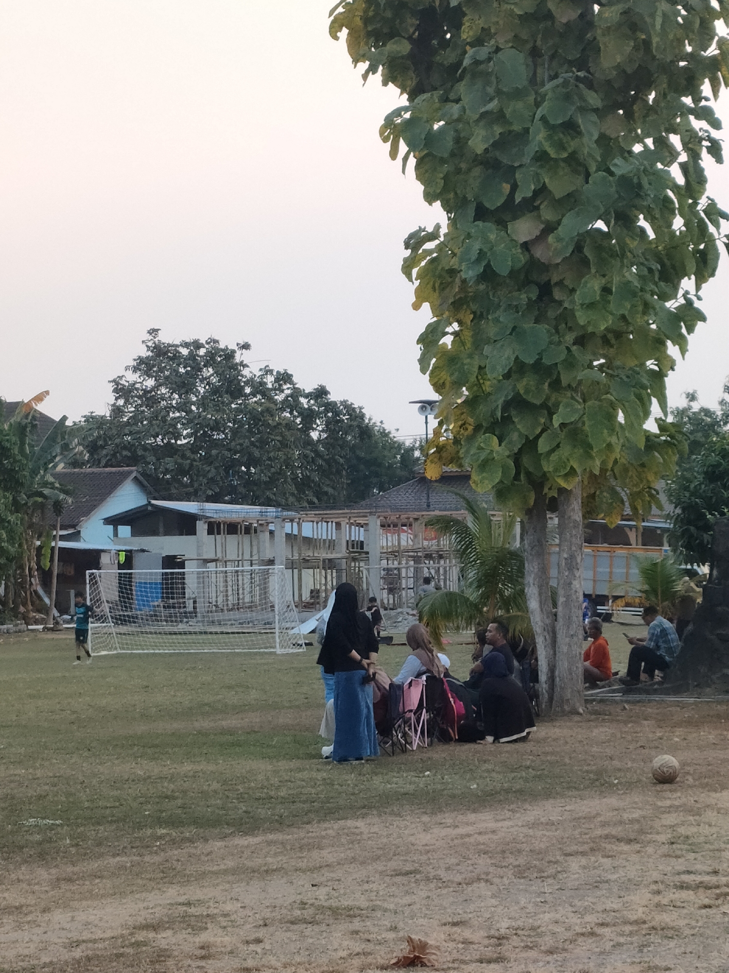 This is a photograph taken outdoors in Banguntapan, Indonesia, depicting a grassy field with a soccer goal in the background. A young person in a blue and black uniform is near the soccer goal. Several adults are seated or standing in groups. One group of people are gathered around what appear to be camping chairs, with one individual wearing a black head covering and a blue skirt. Another person is wearing a pink head covering. A second group of individuals are seated further to the right, including a person wearing an orange shirt and another in a plaid shirt looking at a phone. A large tree with green leaves stands prominently in the foreground, to the right of the center. In the background, there are buildings under construction, some with visible concrete structures and wooden framing. The sky is a pale, hazy color, suggesting it might be late afternoon or early morning. A worn soccer ball is on the ground in the lower right quadrant of the image.