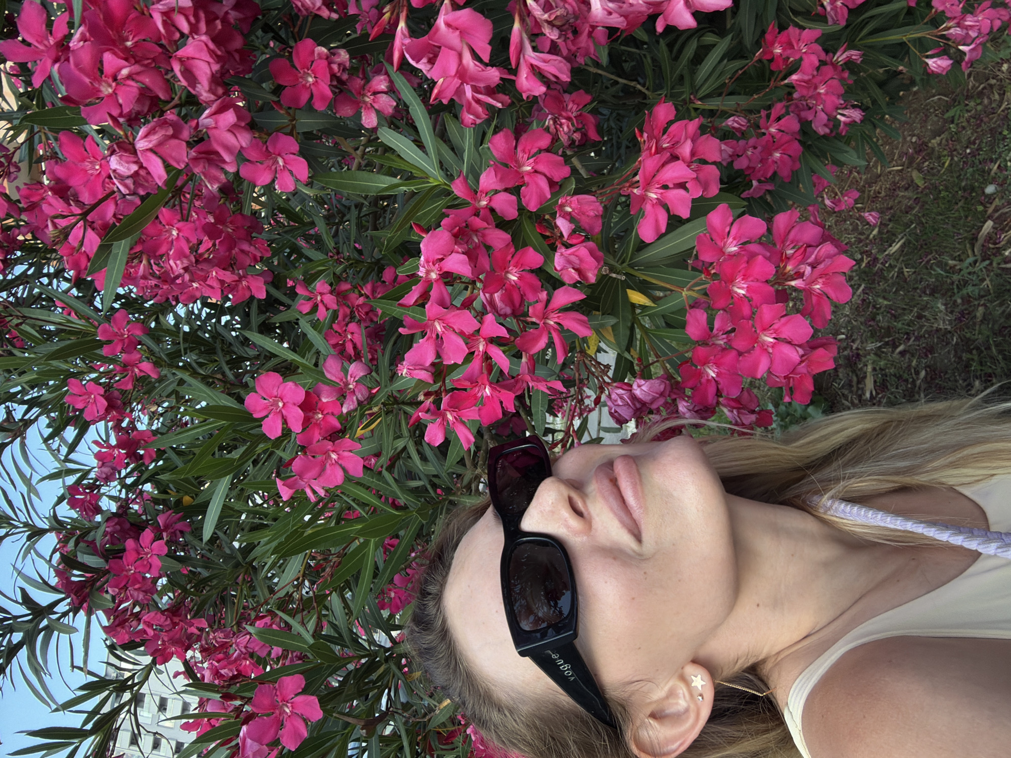 In Budva, Montenegro, a woman with blonde hair and fair skin is pictured from the shoulders up, gazing upwards and to the right. She is wearing large, black, rectangular sunglasses with the word "vogue" printed on the temple, and a light-colored tank top. A delicate gold earring with a star charm hangs from her left ear. She is positioned in front of a lush, dense bush of bright pink oleander flowers. The background reveals a clear blue sky and the corner of a modern, white, multi-story building. The lighting suggests it is daytime, possibly late afternoon, with soft shadows indicating sunlight.
