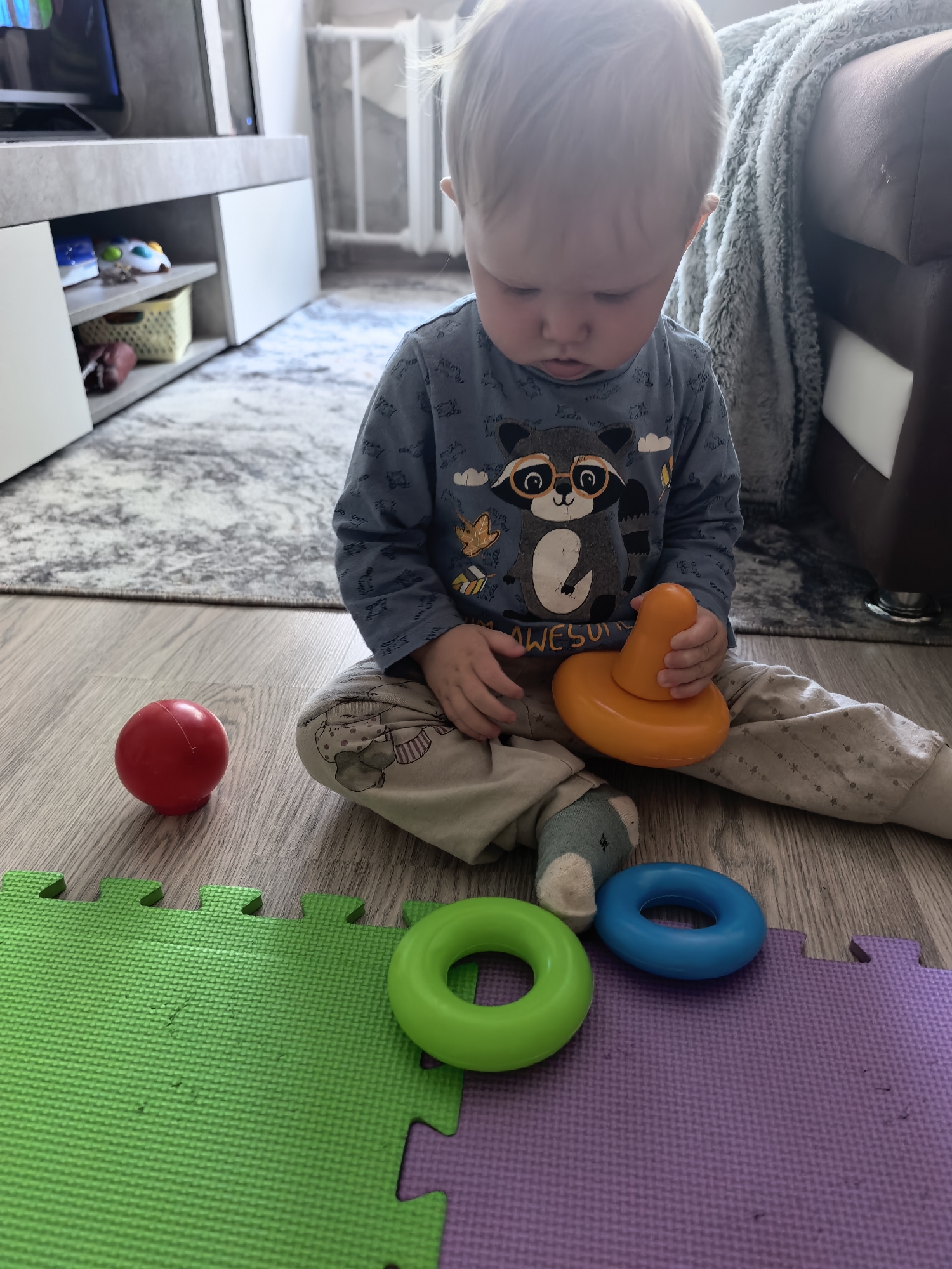 A young child with blonde hair is sitting cross-legged on a light brown laminate floor. The child is wearing a long-sleeved blue shirt with a cartoon raccoon design and light-colored pants with a star pattern. The child is holding an orange toy that appears to be a part of a stacking toy. To the child's left is a red ball. In front of the child are two toy rings, one green and one blue, placed on purple and green interlocking foam play mats. The room contains a light gray entertainment center with white doors, a gray and white patterned rug, and part of a brown couch with a gray throw blanket. The location is Sterlitamak, Russia.