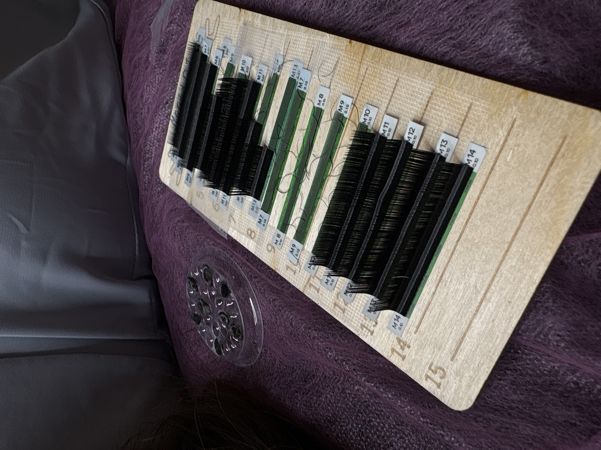 The image shows a wooden lash tray filled with strips of black eyelash extensions, organized by length. The tray is placed on a textured purple fabric, possibly a blanket or shawl. To the left of the tray is a small, clear plastic organizer containing dark circular objects, likely adhesive dots for eyelash application. In the background, a dark gray, draped fabric is visible. The scene appears to be indoors, with soft lighting. No people are visible in the image, and no specific location in Moscow, Russia, can be determined from the visual information provided.