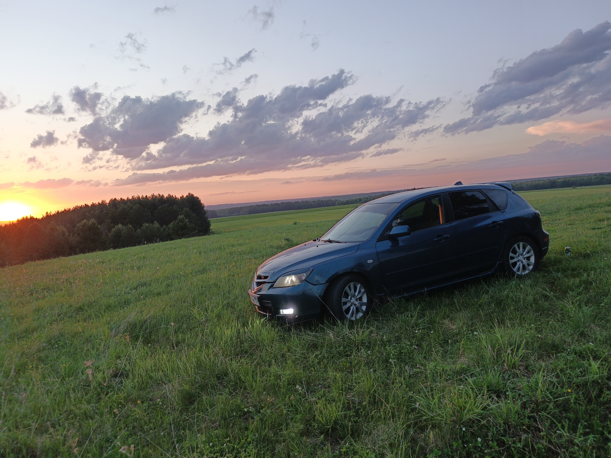A dark blue Mazda 3 hatchback is parked in a grassy field at dusk. The field is located on a slight incline. The sun is setting in the distance over a treeline to the left. The sky is filled with clouds in various shades of orange, pink, and gray. The exact location within Чумна, Russia is not discernible without additional landmarks or context. No people are visible in the image.