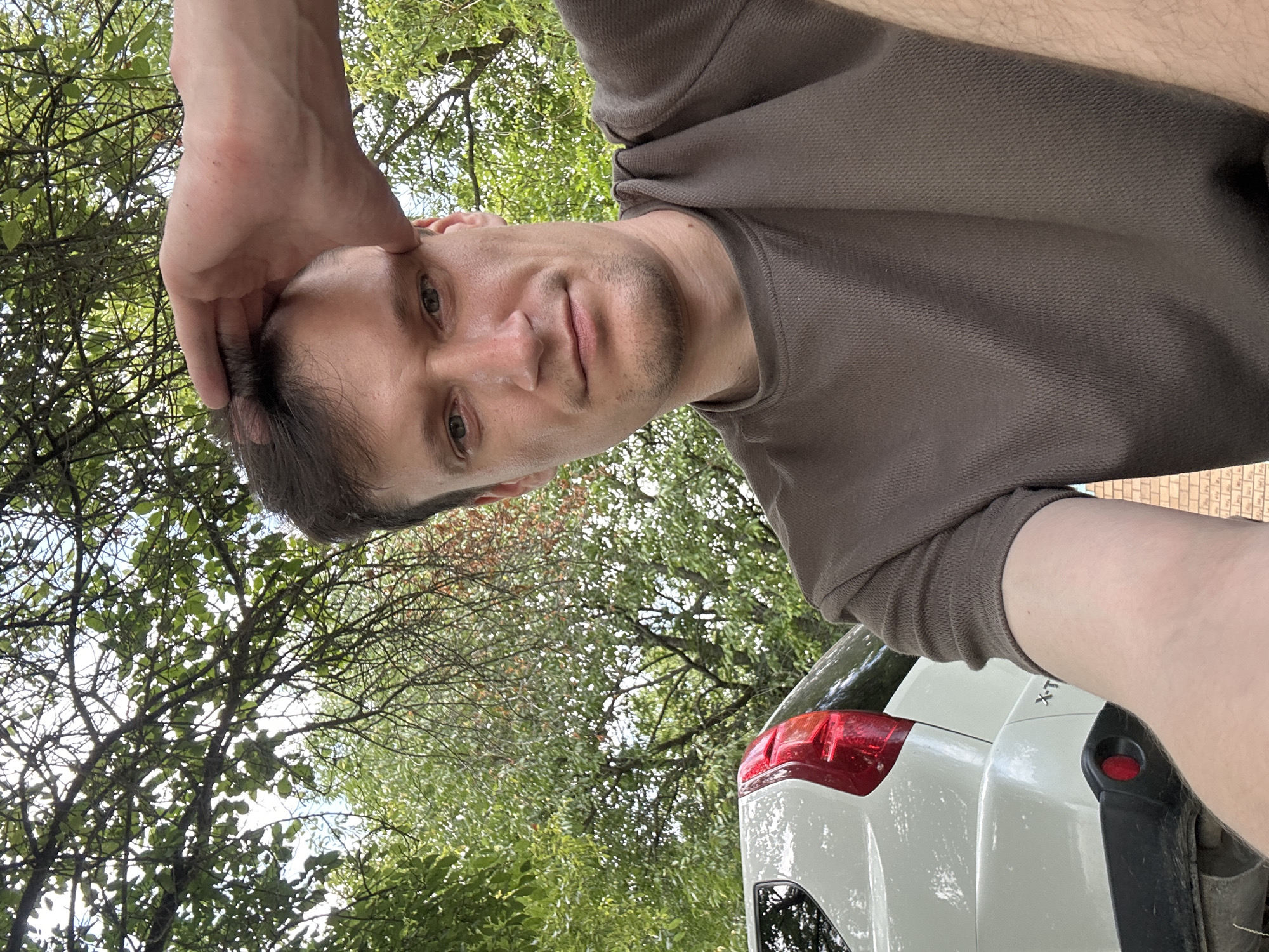 A man is taking a selfie outdoors, with trees and the rear of a white car in the background. He is wearing a brown t-shirt and has his right hand on his head. The car has a red taillight and "X-T" is visible on its bumper. The image was taken in Makiv, Ukraine.