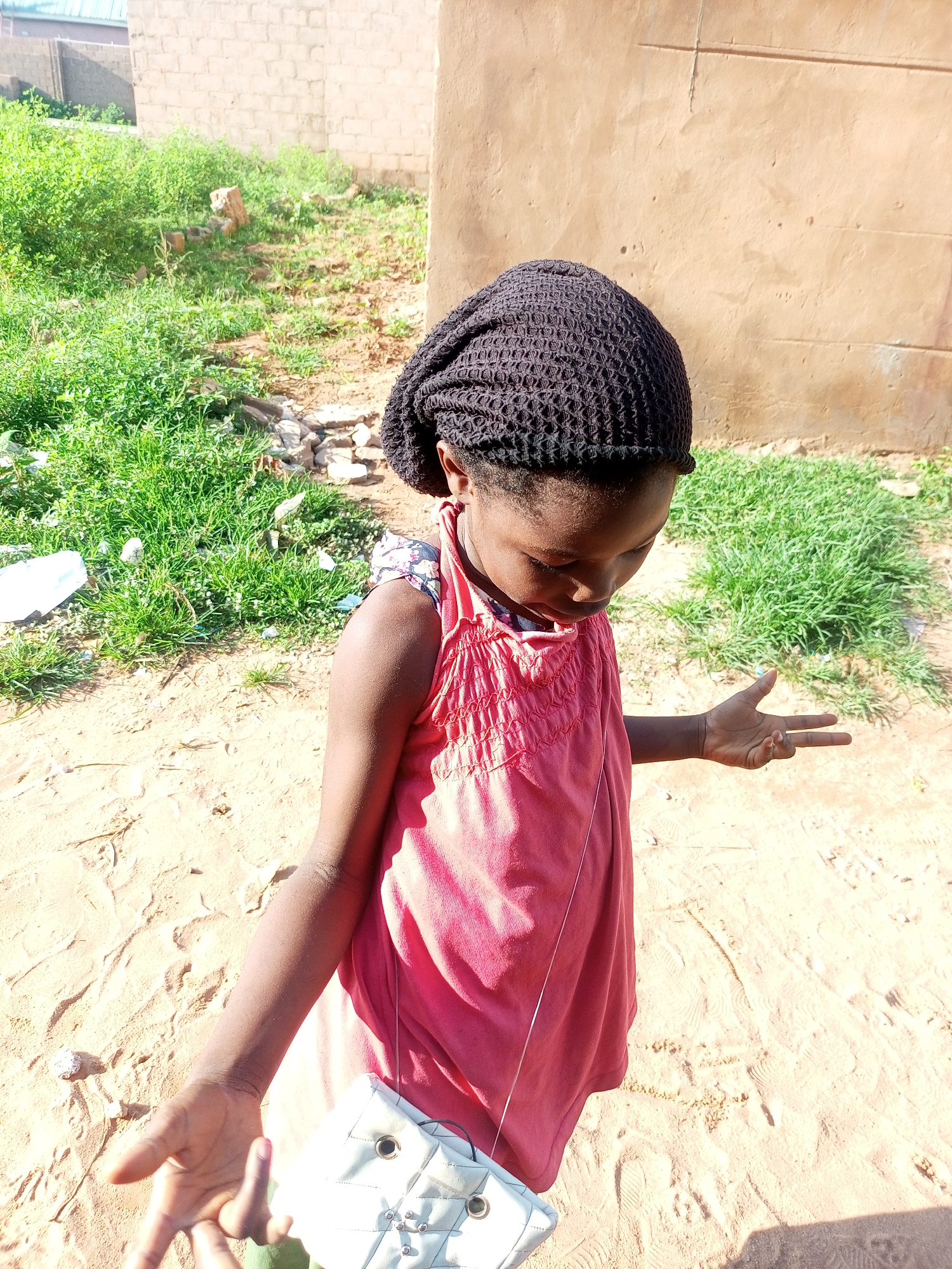 A young girl, appearing to be of African descent, stands outdoors in what seems to be a sandy, unpaved area, possibly within the city of Tempure, Nigeria. She is wearing a dark, knitted cap, a sleeveless pink dress with a patterned insert on the shoulder, and has a light-colored quilted bag slung over her shoulder. The girl is looking downwards and has her left hand raised, with two fingers extended in a V-shape. Her right hand is also extended, palm facing upwards, with her thumb and index finger also forming a V-shape. Behind her, there is a textured, light brown wall and patches of green vegetation, some of which appears overgrown. The ground is sandy with visible footprints and some scattered small stones. Sunlight is casting shadows, indicating it is daytime.