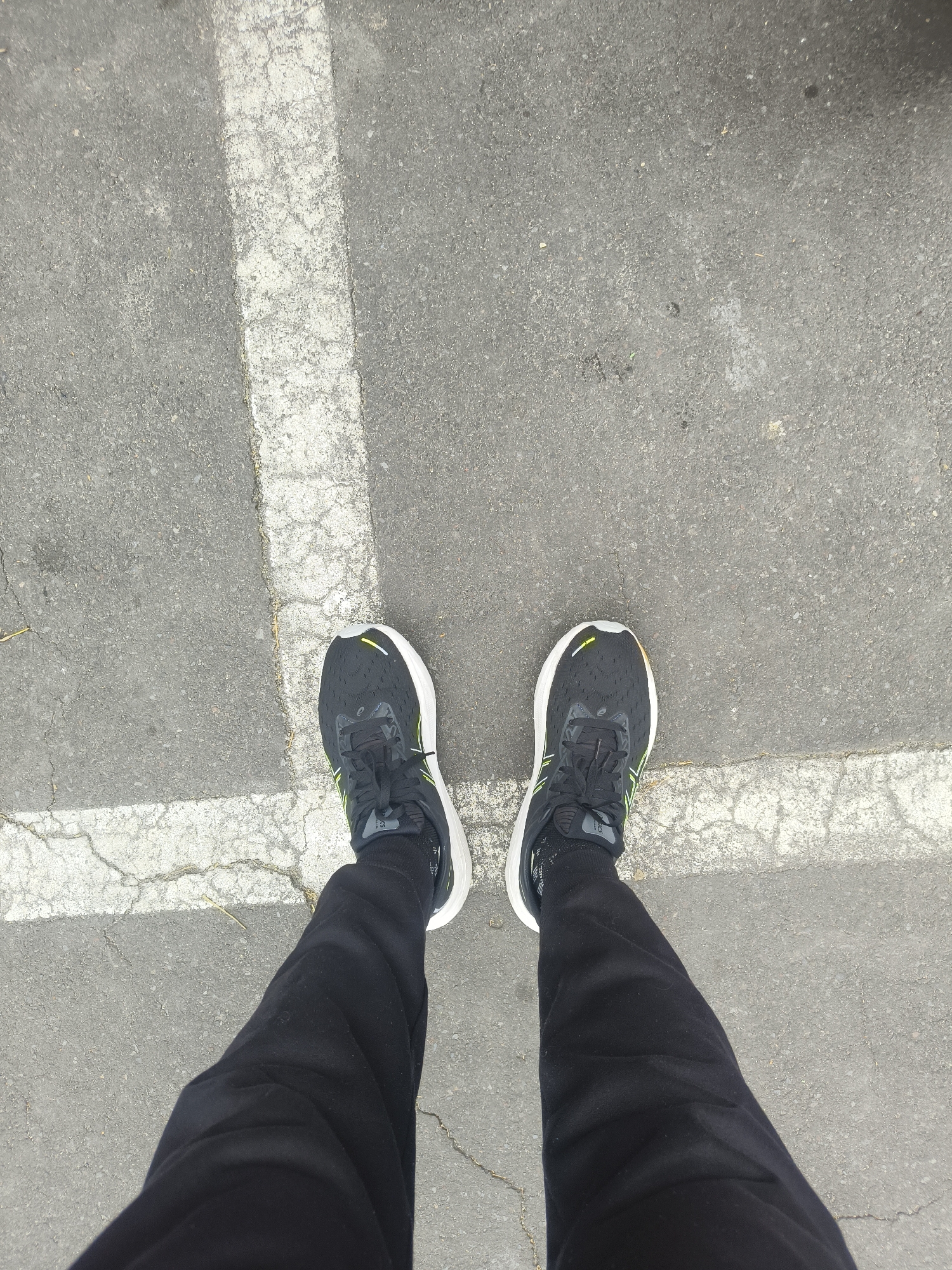 A top-down, full-body shot captures a person standing on asphalt marked with a white line, possibly in a parking lot. The person is wearing black sweatpants and black athletic shoes with white soles and neon green accents. The shoes are tied with black laces. The asphalt is gray and cracked, with some small debris scattered across it. The white line appears to be faded and worn, indicating it has been exposed to the elements. The scene is lit by natural daylight, creating a diffused and overcast atmosphere. The location of Denpasar, Indonesia is not discernible from the visual information provided in the image.