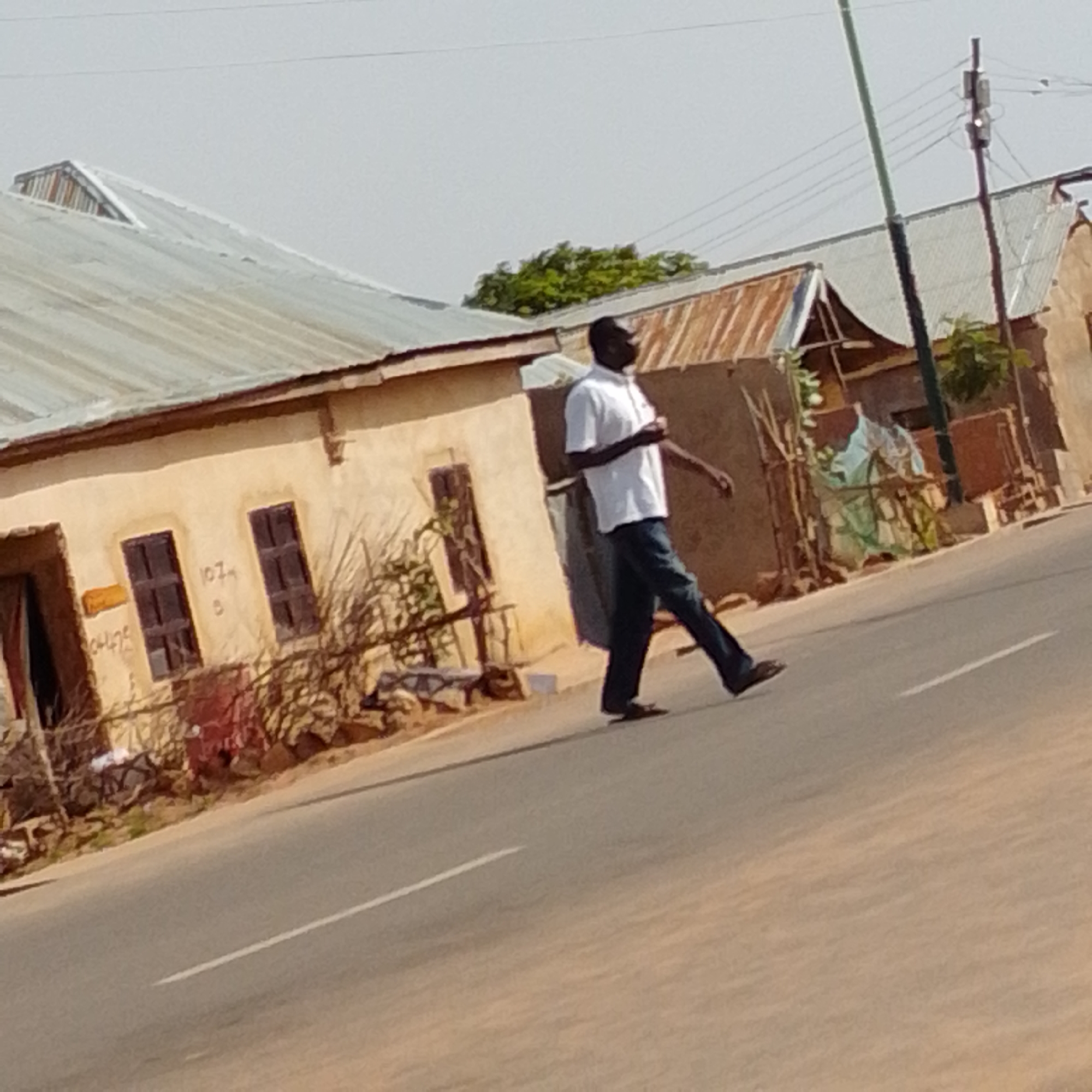 A man in a white shirt and dark pants walks across a street in Gombe, Nigeria. The street is paved with asphalt and has a white line painted in the center. The man is walking from right to left, with his left leg forward and his right leg trailing behind. He is wearing dark shoes.
In the background, there are several buildings with corrugated metal roofs. One building on the left is painted a light tan color and has two dark windows. There is some writing on the wall of this building, but it is difficult to read. Another building on the right appears to be made of mud brick and has some sort of cover made of a light green material. There are some trees and bushes visible in front of the buildings. A tall, thin pole, possibly a utility pole, is also present in the background. The sky is clear and overcast. The overall scene depicts a typical street view in a Nigerian city.