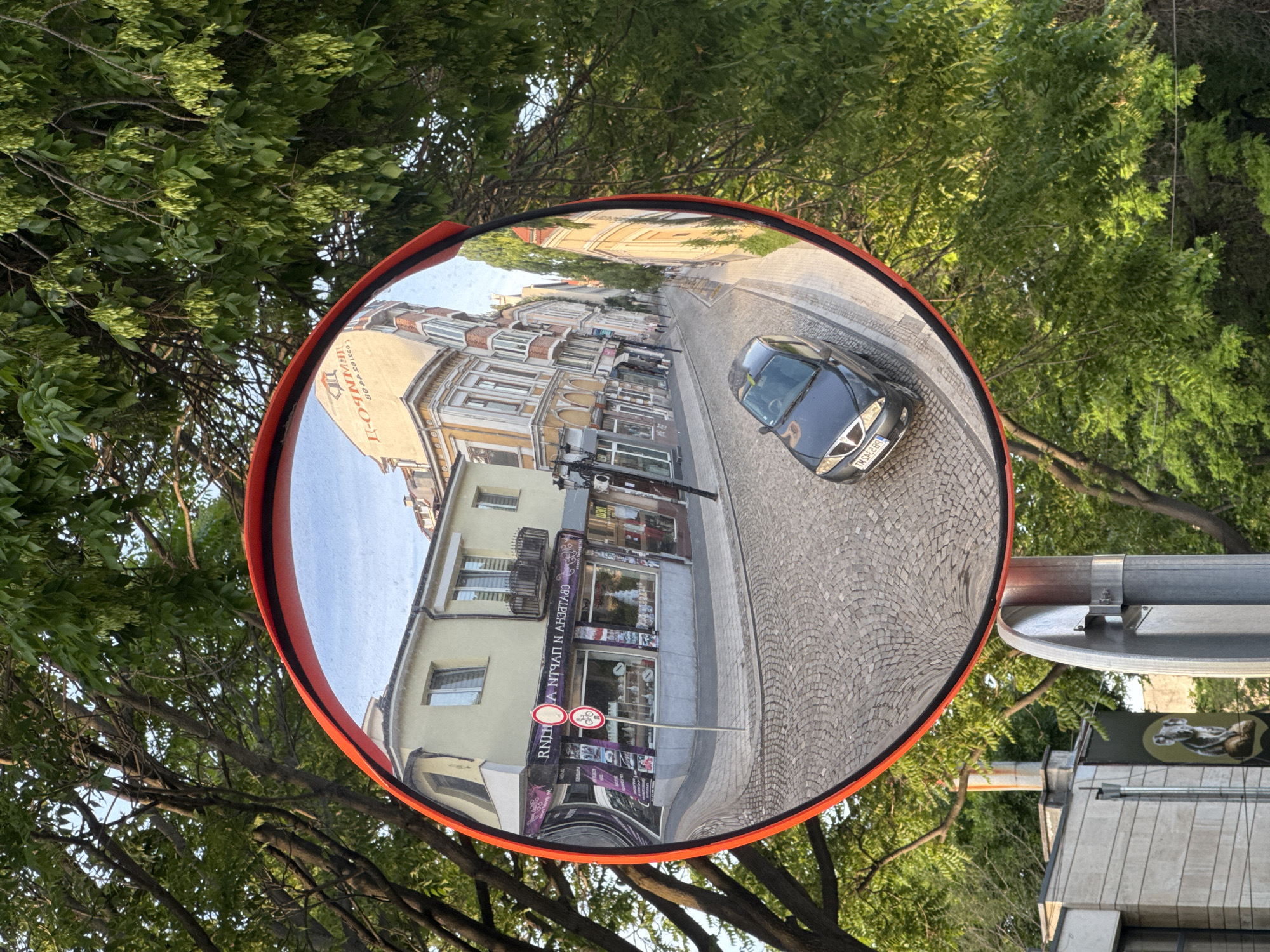 A circular traffic mirror reflects a street scene in Plovdiv, Bulgaria. The reflection shows a narrow, cobblestone street lined with buildings, predominantly two to three stories high. The buildings exhibit a mix of architectural styles with decorative facades and balconies. A dark-colored car is visible driving on the right side of the street. Signage in Cyrillic script and pedestrian crossing signs are visible. The mirror itself has a bright orange border. The mirror is mounted on a gray pole with mounting brackets. Trees with green foliage surround the mirror, partially obscuring the view behind it.
