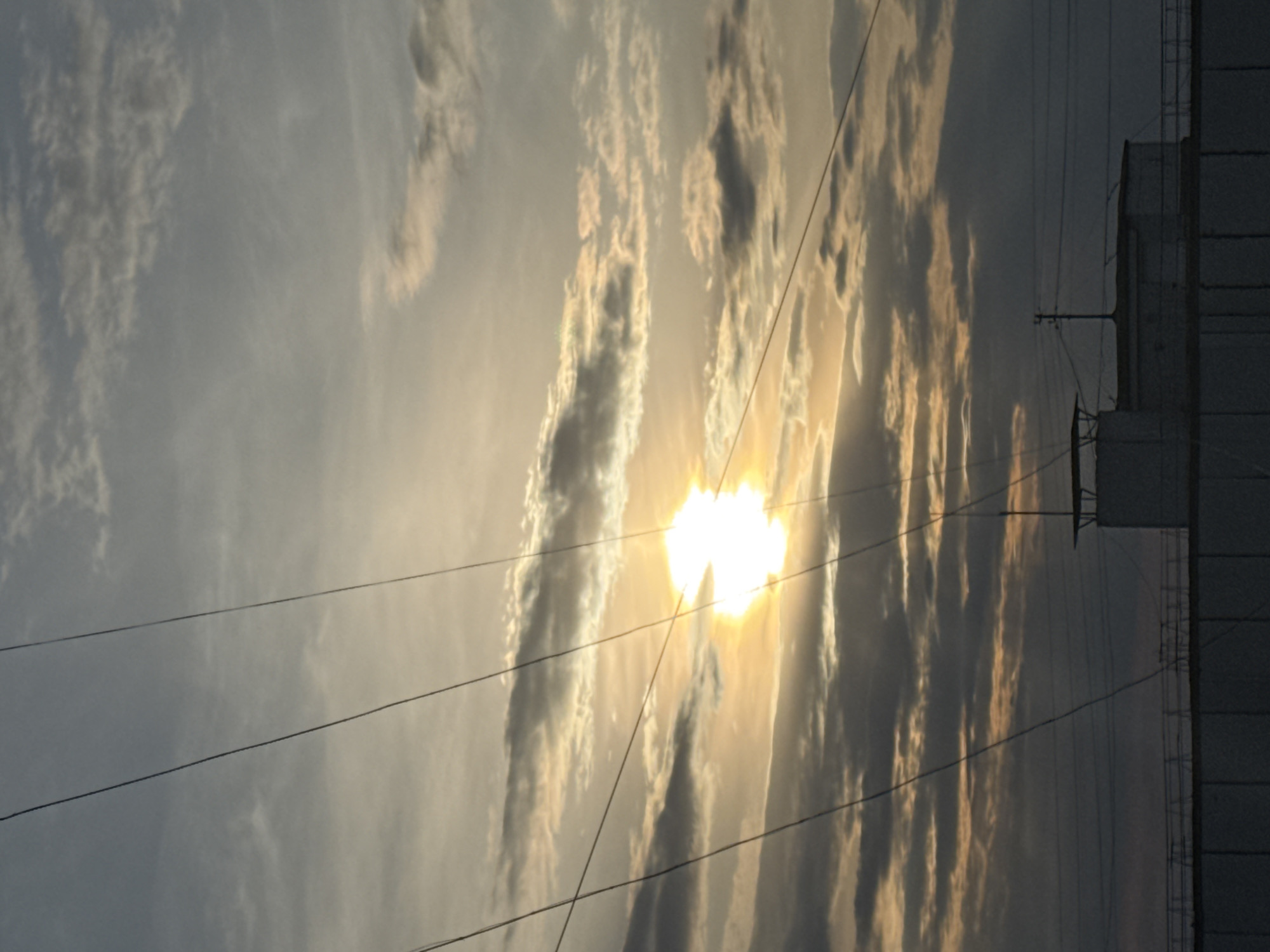 The image depicts a scene in what is likely a city, possibly Chaikovsky, Russia. The view is from the ground looking upwards towards the sky. The sky is filled with clouds and the sun is visible, shining brightly through gaps in the cloud cover. Power lines run across the field of view. On the right side of the image, there is a tall building or structure, likely part of the urban landscape.