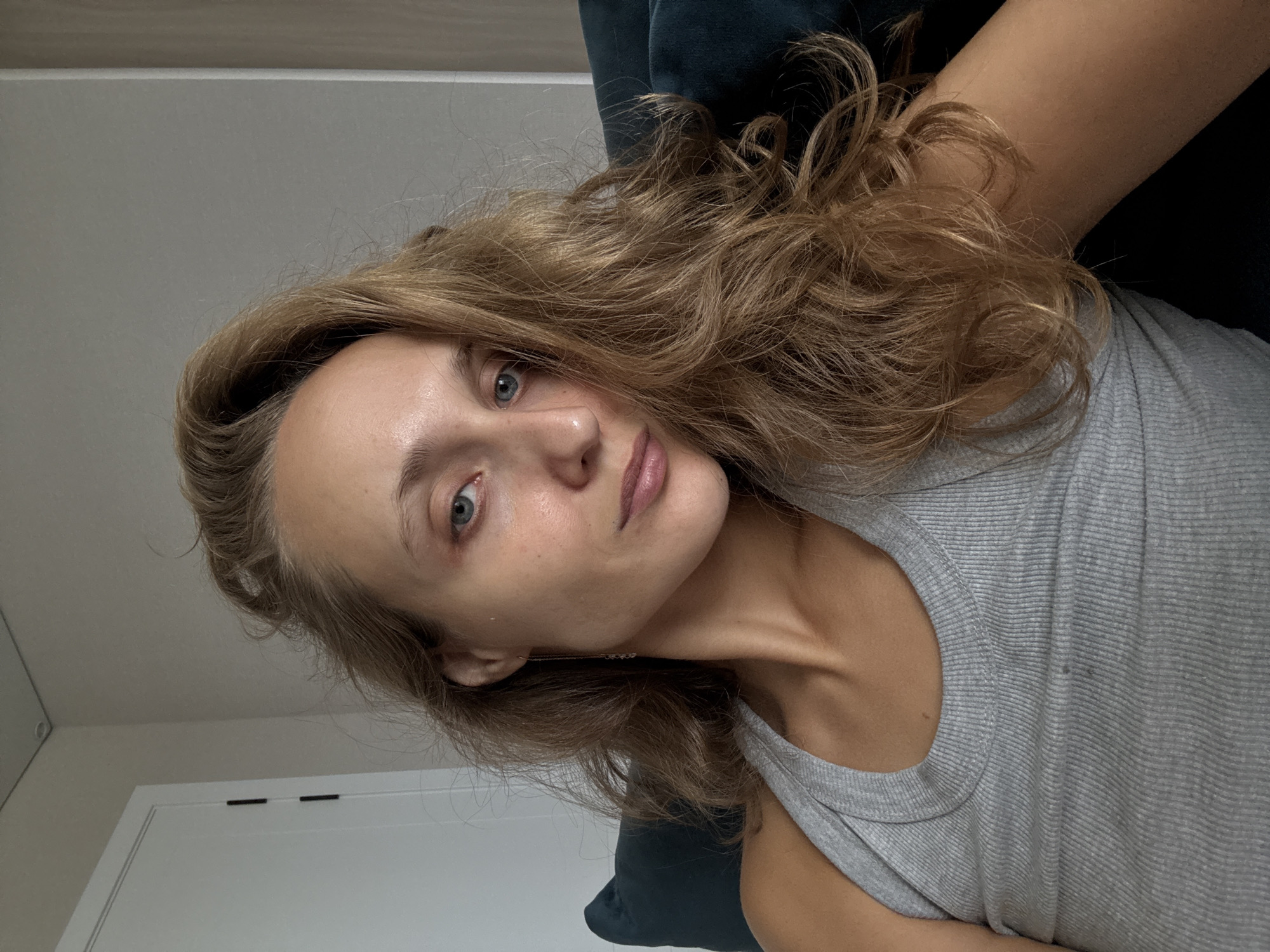 The image shows a close-up, selfie-style shot of a woman with long, wavy blonde hair. She is wearing a grey ribbed tank top. Her head is tilted, and her gaze is directed slightly away from the camera. Her facial features are visible, including blue eyes and light-colored lips. She is seated, with a dark blue cushion visible behind her on the left side of the frame. The background includes a wall and a door on the left. There are no discernible landmarks or visual cues that would indicate a specific location within the city of Подшивалово, Russia, or any other urban environment. The scene appears to be indoors, in what could be a residential setting.