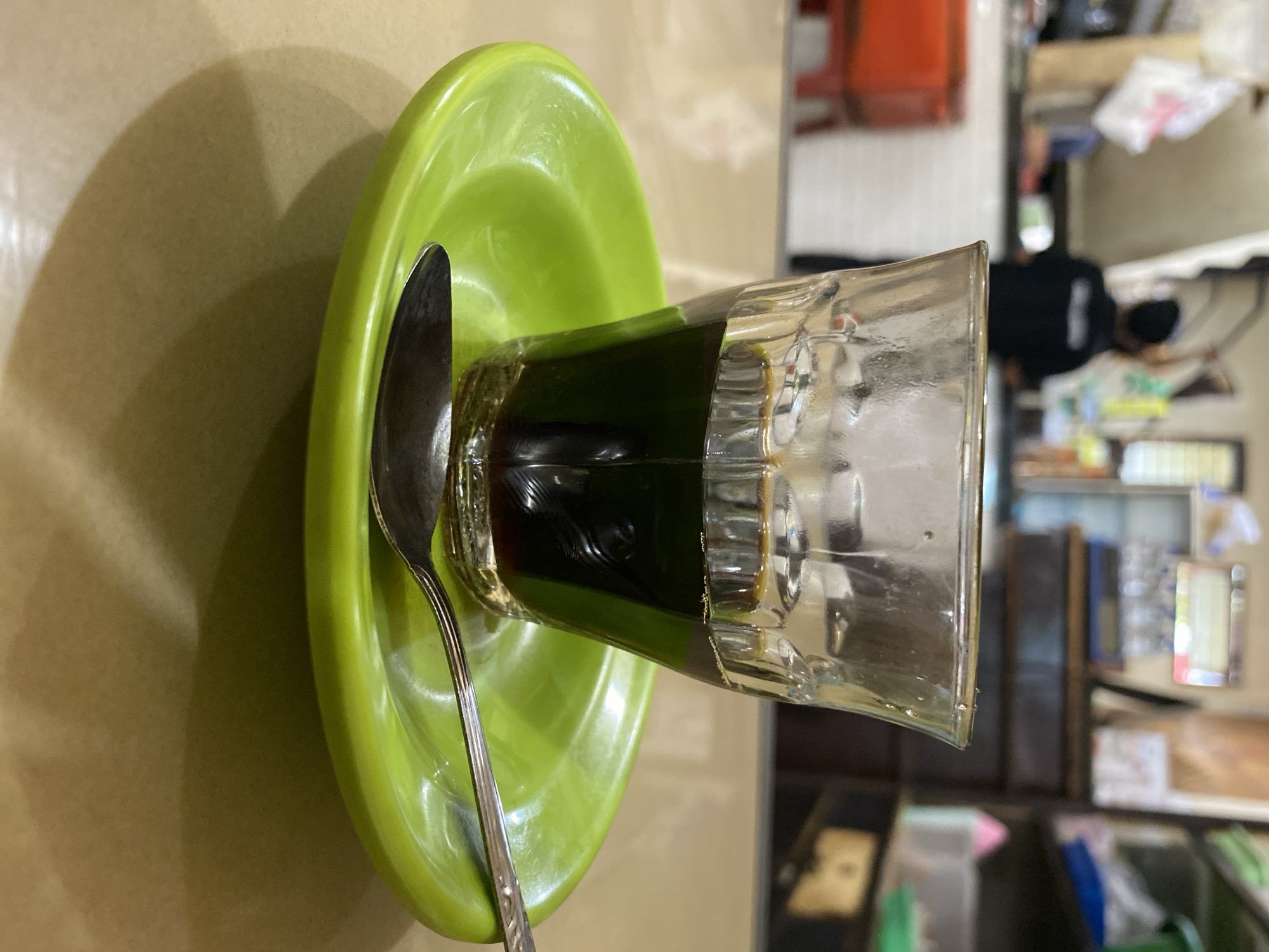 A close-up, eye-level shot captures a small glass of dark liquid, presumably coffee, resting on a vibrant green saucer. A silver spoon lies horizontally on the saucer, positioned to the right of the glass. The glass exhibits condensation and the liquid has a rich, dark hue. The background is blurred, suggesting an indoor setting that appears to be a food establishment or eatery. Hints of shelving units, bags, and indistinct figures are visible, hinting at a busy environment. The table surface is a light brown, textured material. The overall scene suggests a moment of quiet reflection or a pause in activity within a local Indonesian establishment, potentially in the city of Lhokseumawe, Indonesia, given the stylistic elements and common cafe settings found in such regions.