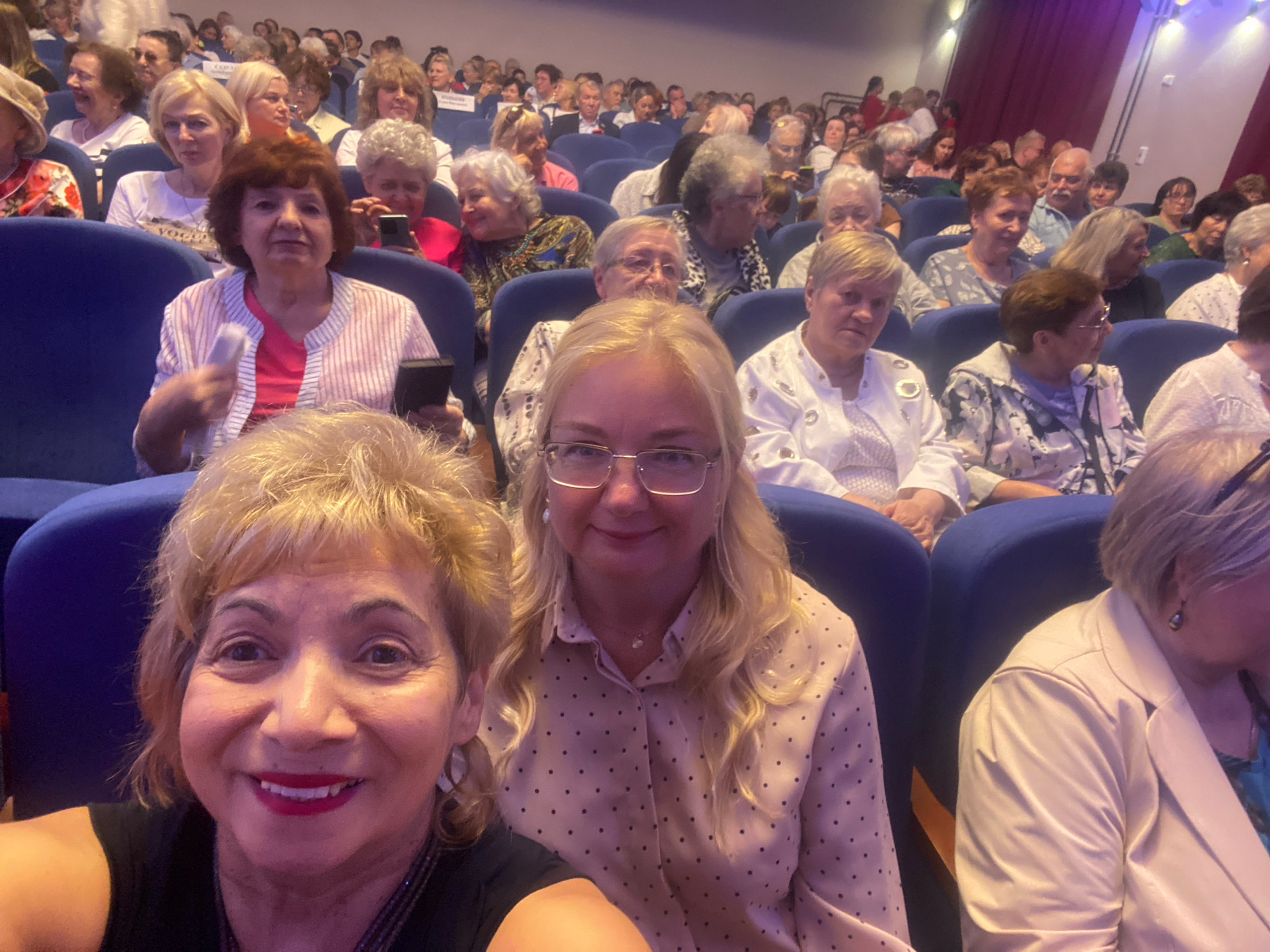 The image shows a group of elderly women attending an event in a theater, possibly a concert or a lecture. They are seated in blue upholstered chairs arranged in rows. The women are dressed in various outfits, with some wearing light-colored blouses and others darker clothing. Several women are smiling, and some are holding phones. The atmosphere appears to be relaxed and enjoyable. The image is taken from a low angle, possibly by one of the attendees. The background shows a stage area with decorative curtains and lighting. The location is specified as Ivanteyevka, Russia, suggesting that this event is taking place in a cultural venue within that city.