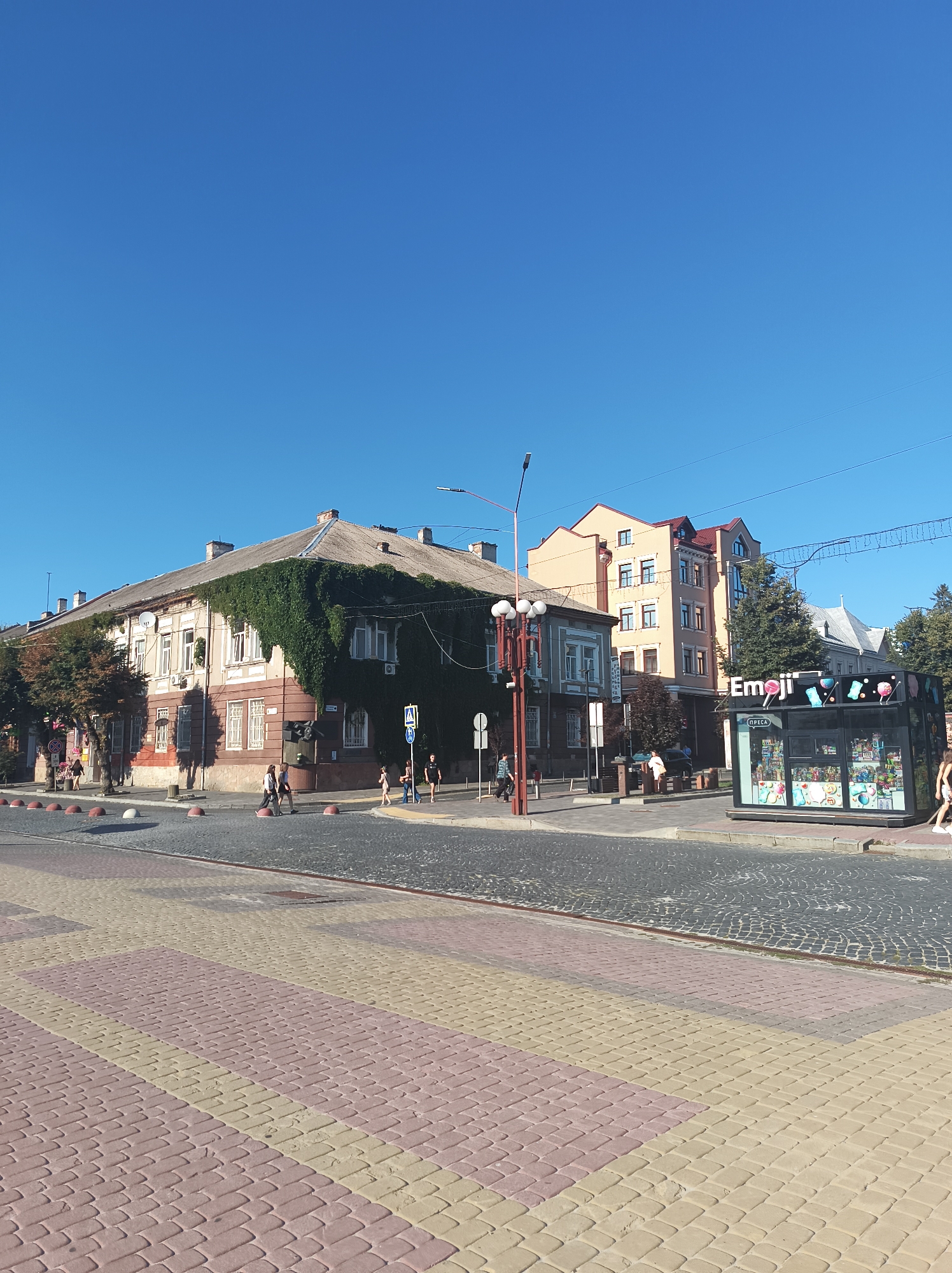 A street scene unfolds under a clear blue sky in Ternopil, Ukraine. In the foreground, a pedestrian walkway is paved with alternating rows of pink and yellow rectangular pavers. This transitions to a cobblestone street, leading to a crosswalk where several people are walking. On the right side of the street, a modern kiosk with the word "Emoji" prominently displayed is filled with colorful merchandise. Behind the kiosk, a multi-story building with a reddish roof stands. To the left, an older, multi-story building is partially covered by dense green ivy, giving it a unique appearance. Several trees line the sidewalk in front of this building, and more buildings of varying architectural styles are visible in the background. A street lamp with multiple lights stands near the ivy-covered building.