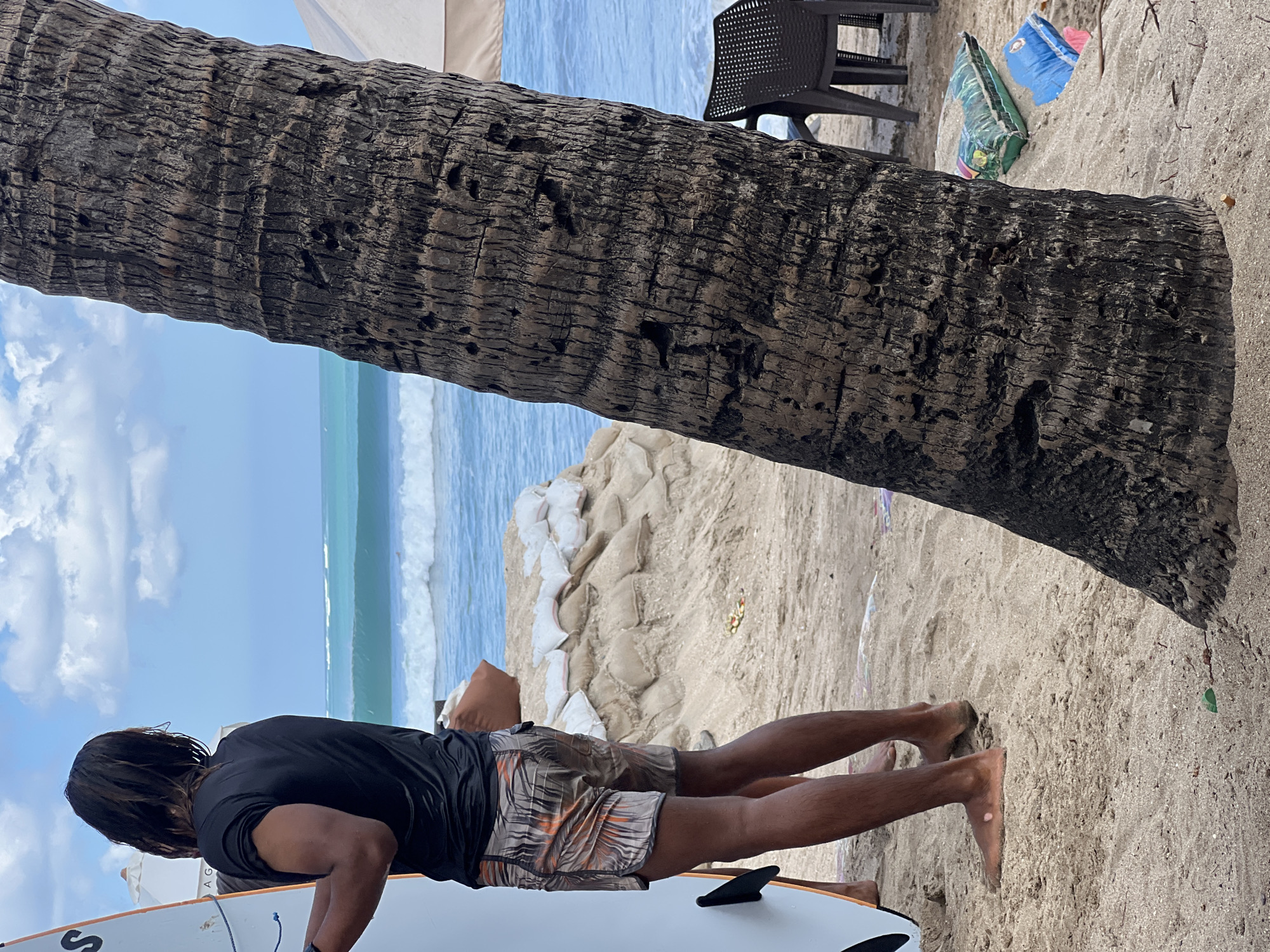 A person stands on a sandy beach in Kuta, Indonesia, holding a surfboard. The person is wearing a black t-shirt and patterned shorts. In the background, waves are crashing on the shore and a palm tree is prominently featured in the foreground. Sandbags are stacked on a sandy embankment behind the person, and beach chairs are visible to the right. The sky is cloudy and blue.