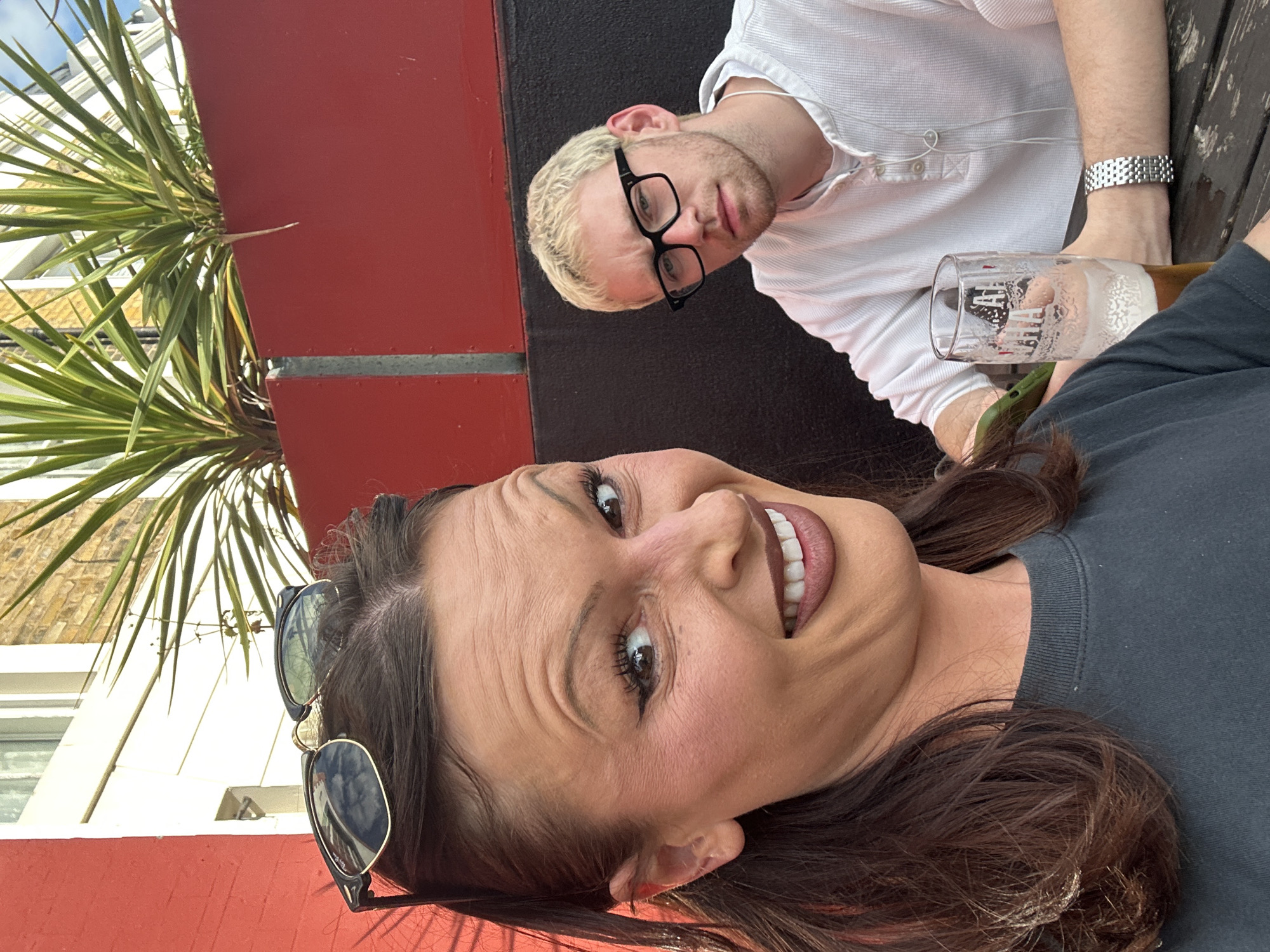 The image shows a selfie taken by a woman with long, dark brown hair and sunglasses perched on her head. She is smiling broadly, displaying her teeth. Next to her, a man with short, blond hair and black-framed glasses is looking at the camera with a neutral expression. He is wearing a white, button-down shirt and a silver watch on his left wrist. A pint glass filled with beer is visible in front of him.
The background indicates an outdoor setting, likely a pub or bar. A red-painted structure, potentially a wall or planter, is visible behind the people, and green, leafy plants are growing nearby. Given the architectural details faintly visible in the background, this setting is likely located in the city of London, United Kingdom.