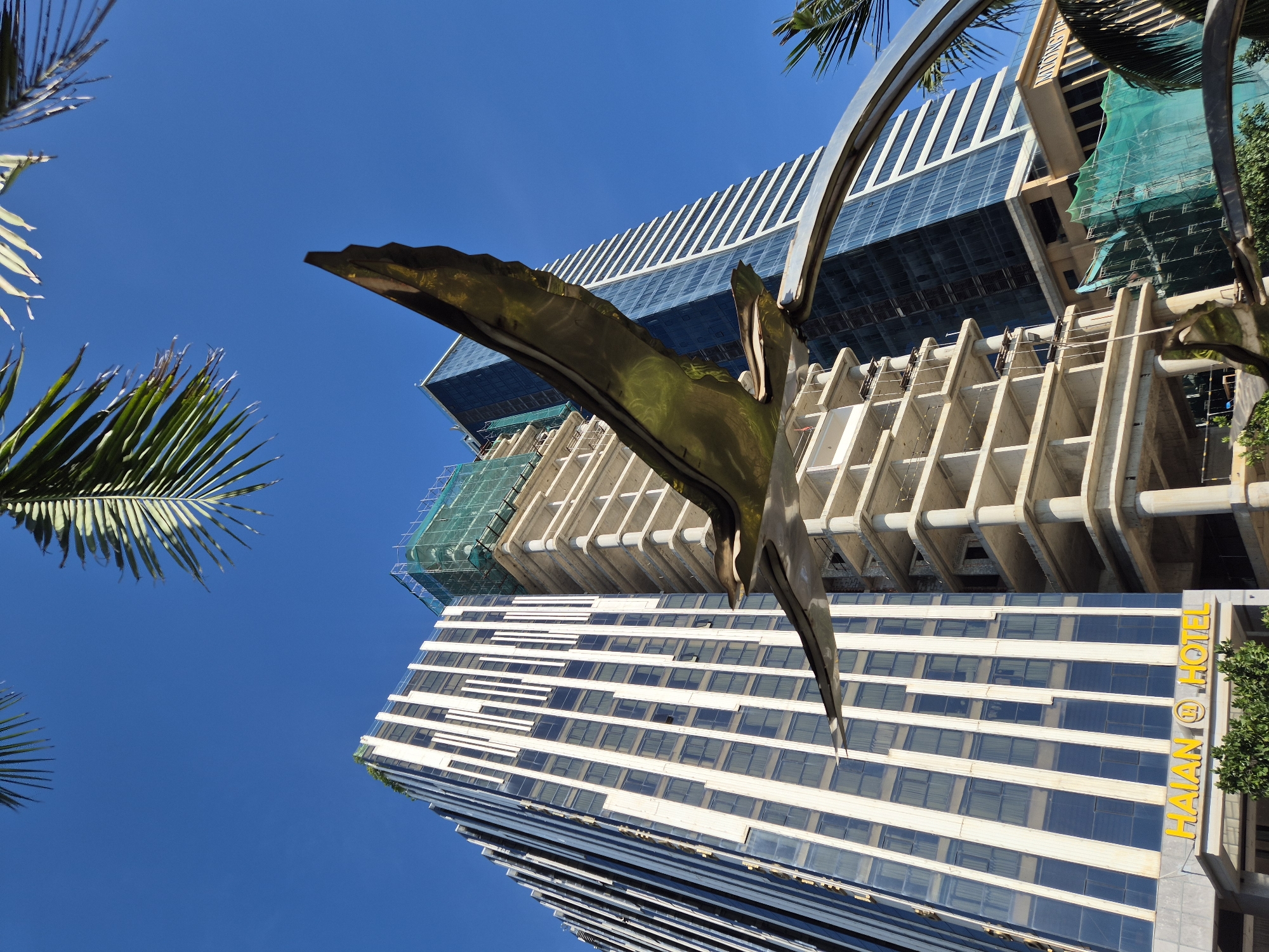 A low-angle outdoor photograph captures a sculpture of a bird in mid-flight in front of several high-rise buildings under a clear blue sky in Đà Nẵng, Vietnam. The sculpture, made of a reflective metallic material, is positioned in the foreground, with its wings outstretched and its head facing towards the right of the frame. The bird sculpture appears to be a focal point of a public art installation.
Behind the sculpture, a modern hotel building, identified by a prominent "HAIAN HOTEL" sign with a circular logo, stands to the left. This building features a facade of alternating dark glass windows and white vertical panels. Adjacent to the hotel, a building is under construction, with its concrete structure exposed and scaffolding and green netting covering parts of its exterior. Further to the right, another glass-fronted skyscraper rises, reflecting the blue sky.
Palm fronds frame the top of the image, suggesting a tropical or subtropical location. The scene depicts an urban environment with new development, characterized by the juxtaposition of established architecture, ongoing construction, and public art. The lighting indicates it is daytime, with the sun illuminating the buildings and sculpture.