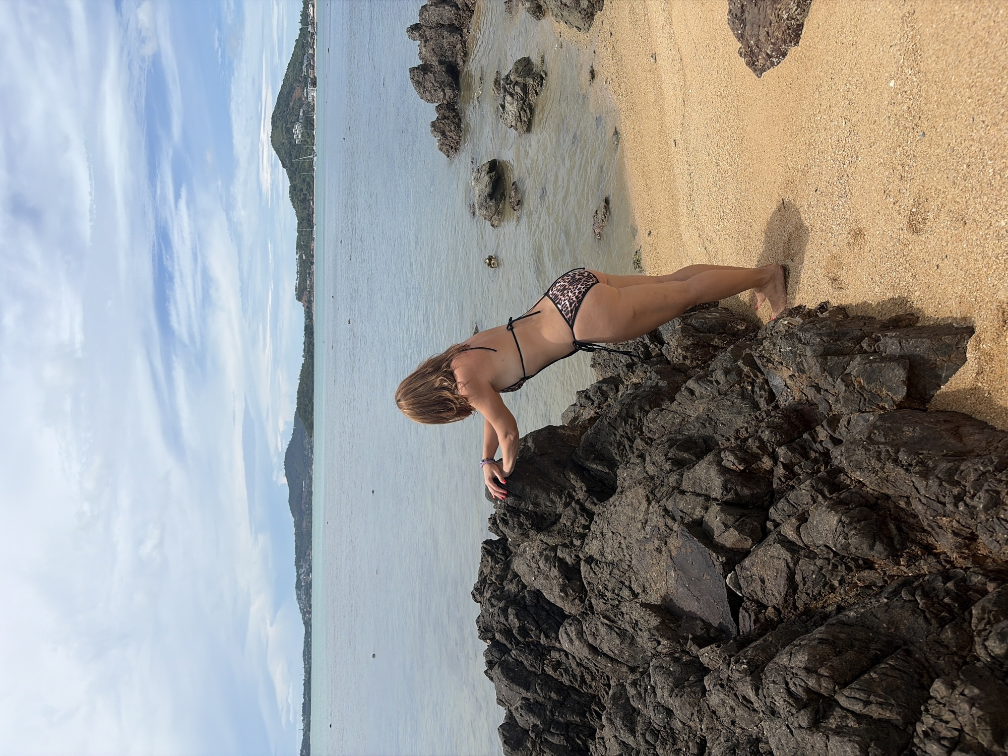 A woman in a leopard-print bikini leans over dark, rough rocks on a sandy beach in Baan Bo Phut, Thailand. She is facing away from the camera, towards the ocean. The water is calm and light blue, with several small rocks visible near the shore. In the distance, a line of green hills with buildings dotted across them forms the horizon. The sky is a pale blue with wispy white clouds. The sand is light brown and appears to be fine-grained.