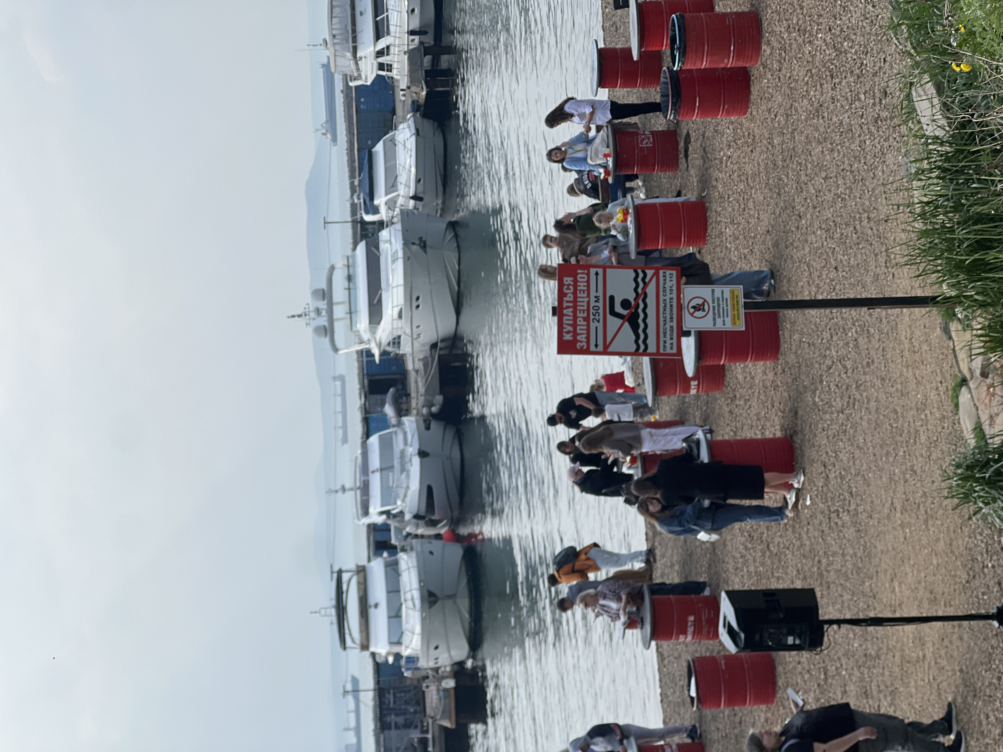 The image shows a waterfront scene in Vladivostok, Russia. Several small white boats are docked on the left side of the image next to a calm body of water, likely the sea. On the right side of the image, a row of red barrels lines a gravelly area next to grass. People are sitting on or standing near the barrels. A sign prohibits swimming, with Russian text indicating "КУПАТЬСЯ ЗАПРЕЩЕНО! - 250 м" (Swimming is prohibited! - 250m). There is also a secondary sign with a person walking their dog, with text underneath reading, "РАЗВЕДЕНИЕ КОСТРОВ." A black speaker on a stand is visible near the barrels, and the sky is overcast.