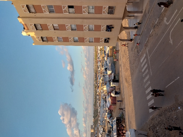 This image captures an outdoor scene showcasing a blend of urban architecture and a densely populated informal settlement or camp, all under a clear sky. The perspective is from an elevated vantage point, looking down upon the scene.
On the left, a multi-story, light brown building with decorative white patterns around its windows dominates the foreground. Several windows are visible, some appearing open, with clothes hanging from a few. Below the building, people are gathered, walking, and standing on a dirt path.
A paved road cuts diagonally through the middle-right of the frame, featuring faded white markings, including a visible crosswalk. A small group of people is standing on the crosswalk, while other individuals are scattered along the road and its dusty shoulders.
Extending from the right side of the road into the background is a vast area filled with numerous informal structures, tents, and makeshift shelters, characteristic of a crowded camp or informal settlement. Many more people can be seen moving about within this area.
The sky above is clear blue with several scattered white and light gray clouds. The lighting suggests either late afternoon or early morning, with long shadows stretching across the ground and a warm light illuminating parts of the building, indicative of a low sun angle. No discernible text is visible on any signs or objects. The scene depicts everyday life in an environment that combines permanent structures with a sprawling, informal dwelling area, consistent with the context of Khan Yunis, Palestinian Territory.