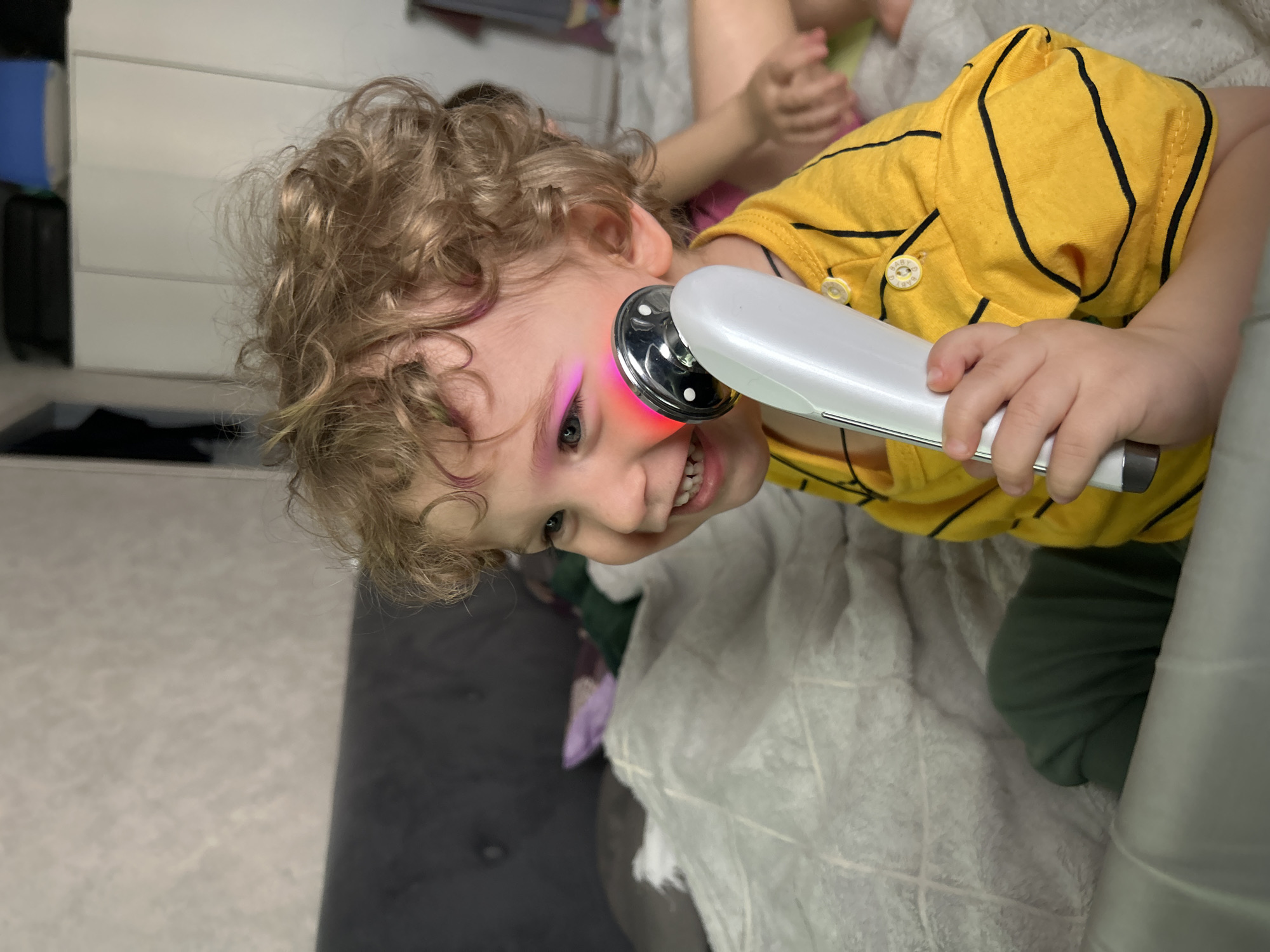 A child with blond curly hair is holding a white and silver device with a red light against their face. The child is wearing a yellow and black striped shirt and appears to be lying on a bed or couch with a white patterned blanket. Another child's hands are visible in the background. The location is Klin, Russia.