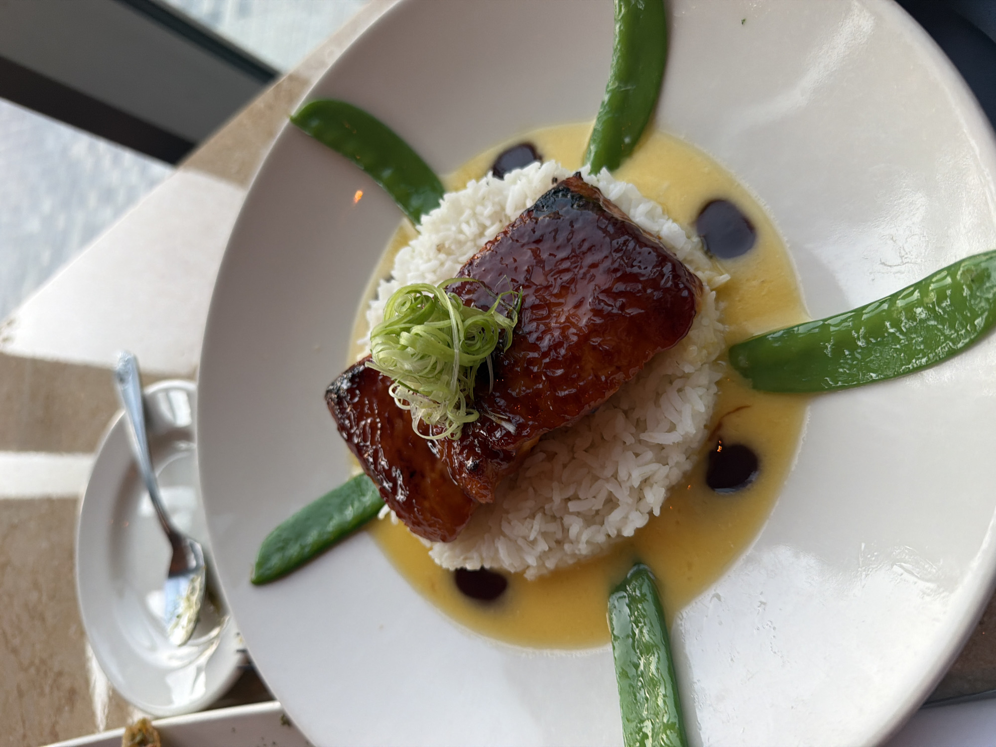 A close-up, eye-level shot captures a beautifully presented dish on a white plate. The dish features a mound of white rice topped with two pieces of glazed, dark-brown protein, possibly fish or duck, glistening under the light. A garnish of finely chopped green onions sits atop the protein. The plate is surrounded by a light yellow sauce, with several bright green snap peas arranged around the perimeter. Two dark, circular elements, possibly a reduction or sauce, are visible within the yellow sauce. The plate rests on a table with a textured, beige surface, and in the background, a glimpse of a window reveals a blurry, bright outdoor scene, suggesting an elevated position with a city view. A small, white saucer with a silver spoon is partially visible on the left side of the frame, indicating this is likely a restaurant setting. The lighting is bright, highlighting the textures and colors of the food. There are no people visible in the image, and no specific details are visible to indicate a location in Dubai, United Arab Emirates.